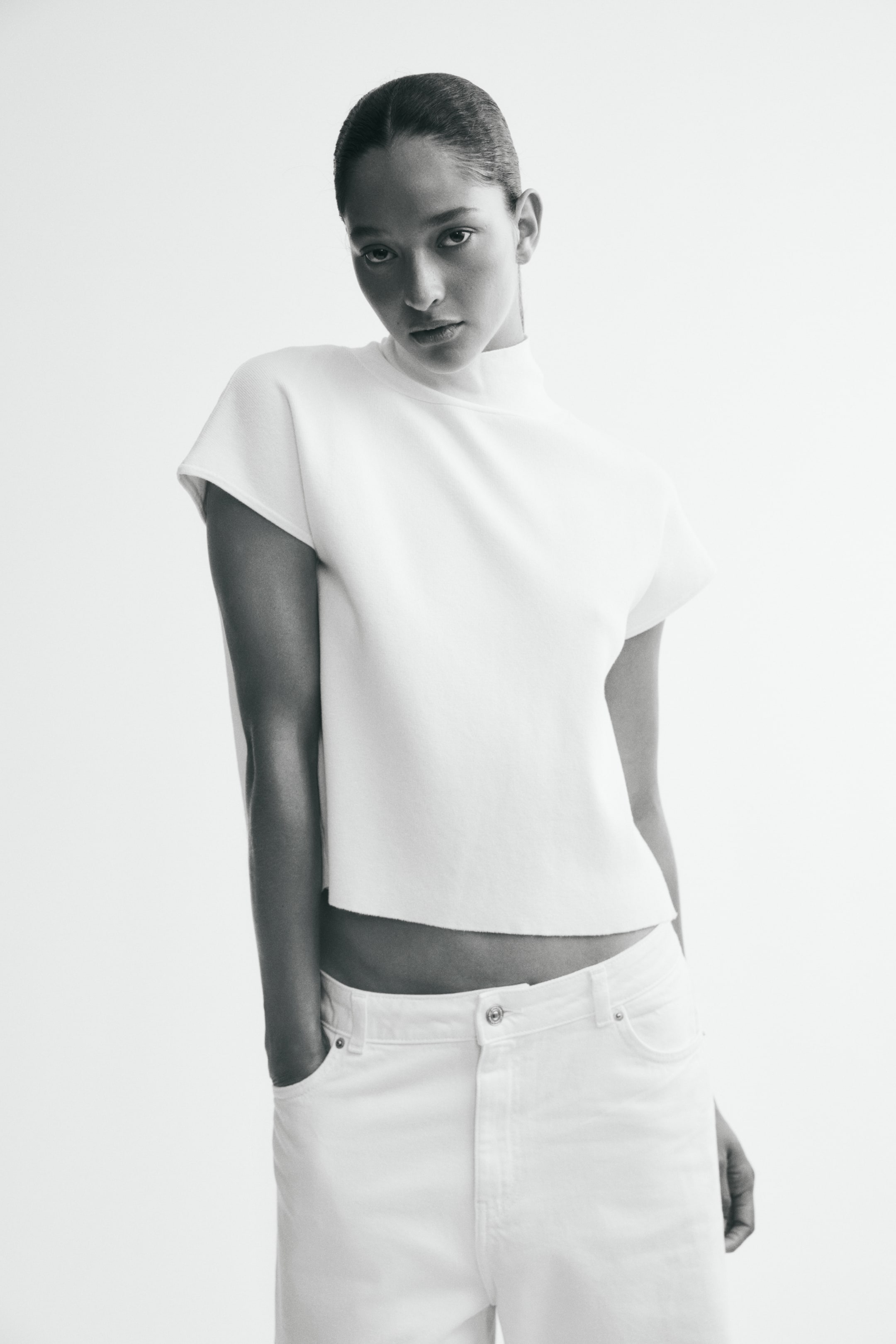 Ver imagem maior: Um top branco cropped apresenta uma gola alta, mangas curtas e uma bainha reta com acabamento cru. É combinado com jeans brancos de cintura alta, que exibem bolsos frontais e fecho de botão.