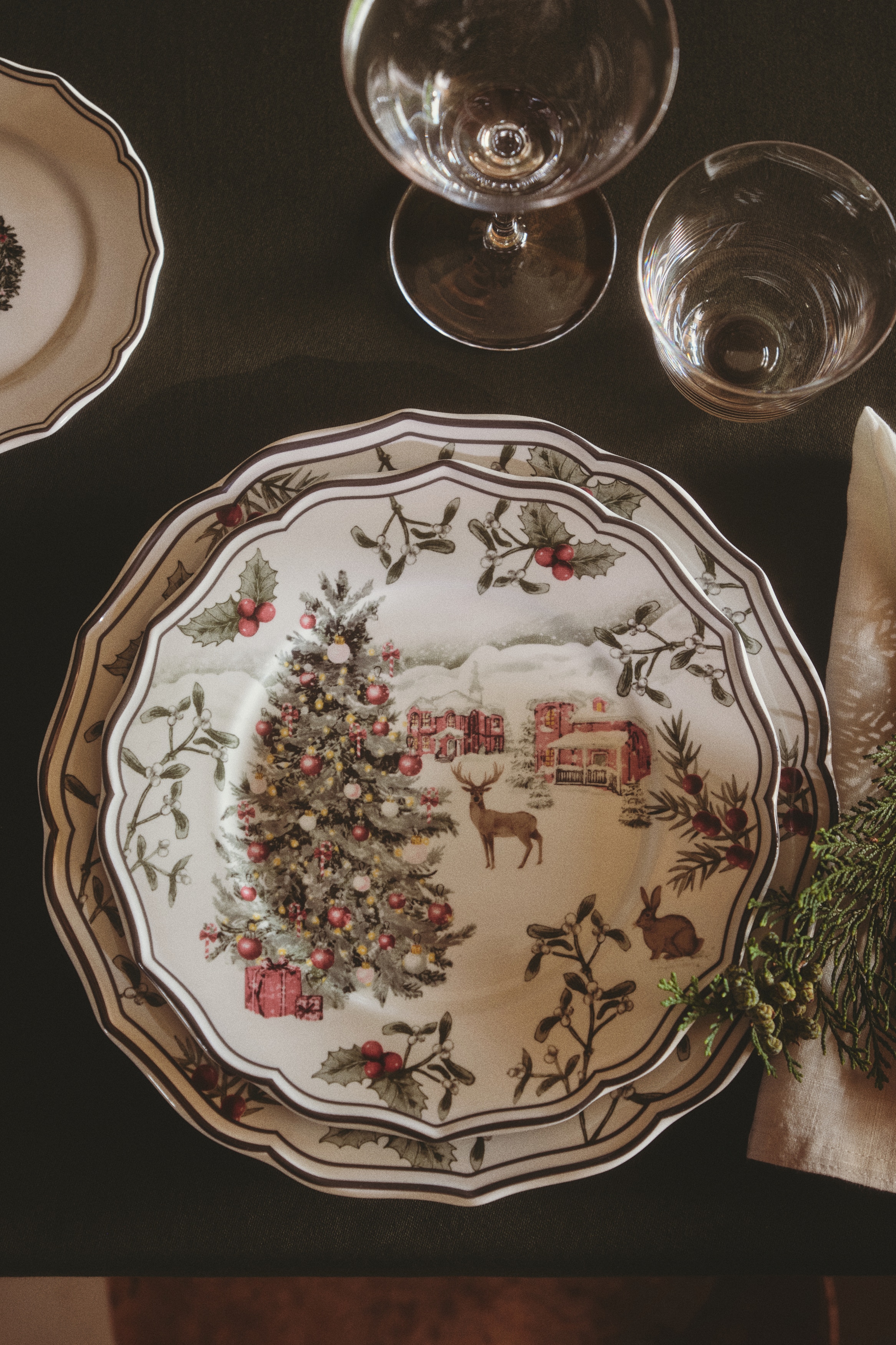 A set of four white ceramic plates is displayed from a front-facing view in a two-by-two arrangement. Each plate features a scalloped edge with a dark rim and showcases a winter scene illustration that includes a decorated holiday tree with gifts, a deer, and a pink cottage in a snowy landscape.