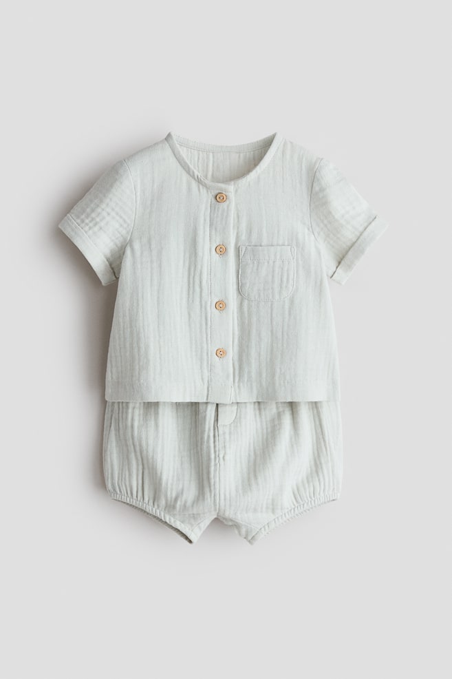 A cotton muslin shirt and shorts set is showcased from the front. The shirt features a round neckline, a button-down front with four light-colored buttons, an open chest pocket, and short sleeves with sewn-in turn-ups. The shorts come with an elasticated waist, two decorative buttons at the front, and narrow elastication around the legs. - 1