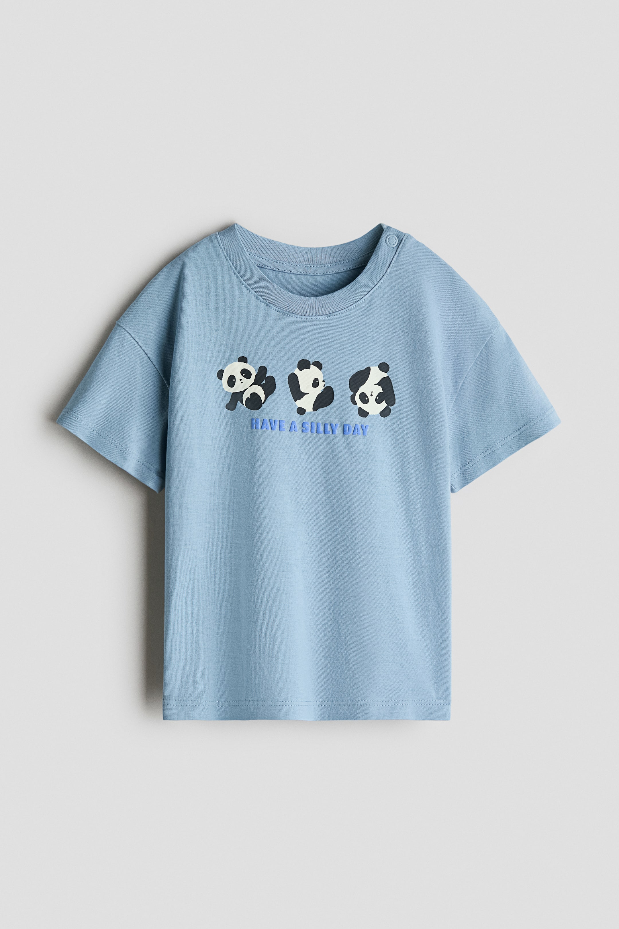 View larger image: A printed cotton T-shirt displays its light blue front with a ribbed crew neck and short sleeves. Three cartoon pandas are printed across the chest, with the text "HAVE A SILLY DAY" appearing below them. A single snap button is visible on the left shoulder.