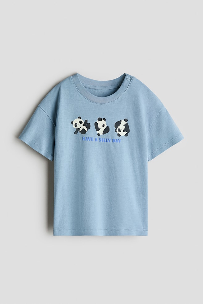 A printed cotton T-shirt is showing its light blue front with a ribbed crew neck and short sleeves. Three cartoon pandas are printed across the chest, and the text "HAVE A SILLY DAY" is appearing below them. A single snap button is visible on the left shoulder. - 1