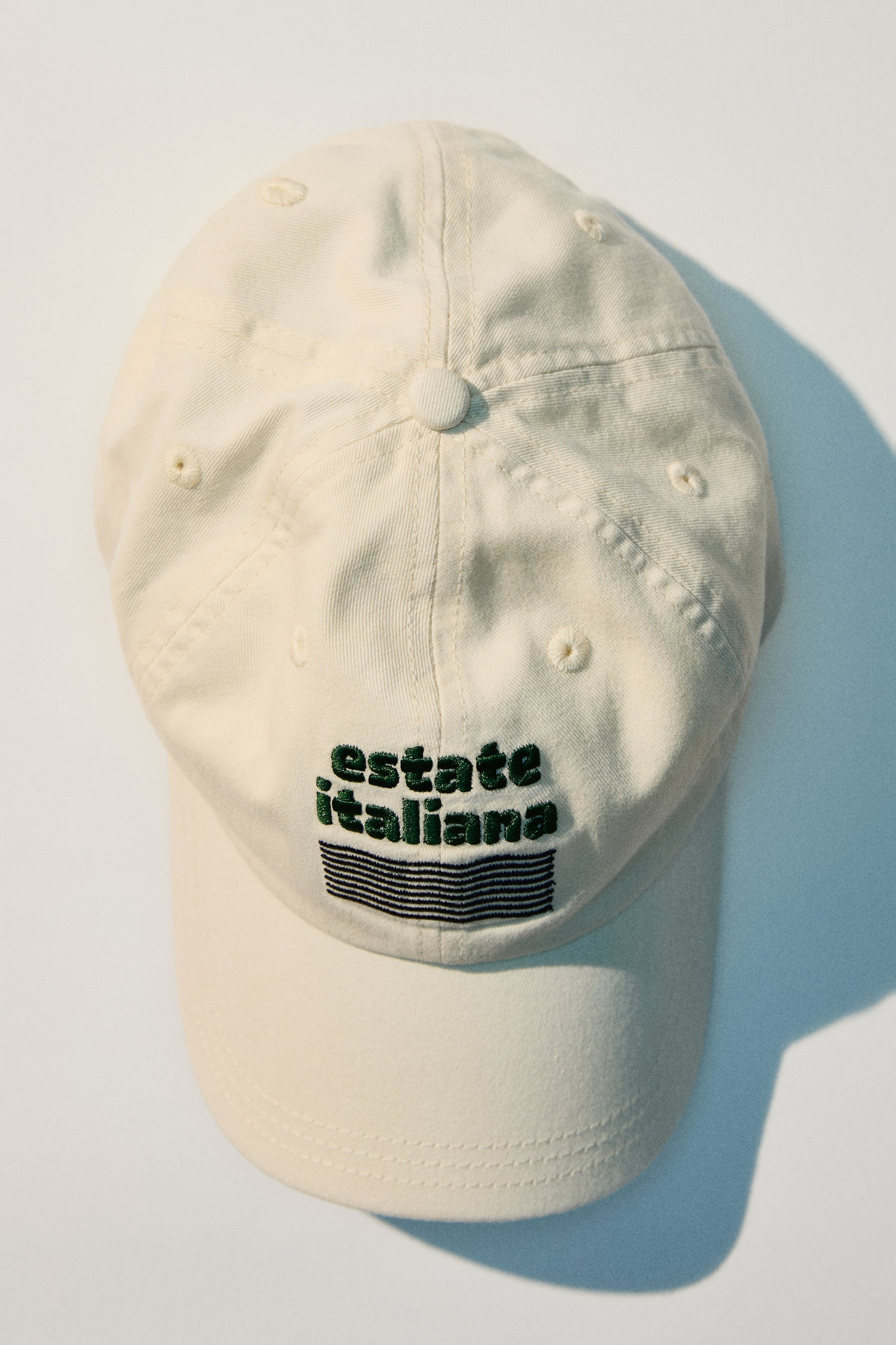 Agrandir l'image: La casquette de baseball de couleur crème est légèrement inclinée d'en haut, orientée vers l'avant, et présente un bouton rond sur sa couronne ainsi que des œillets cousus. Son panneau avant affiche le texte brodé en vert "estate italiana" avec des rayures horizontales noires, et est complété par une visière incurvée.