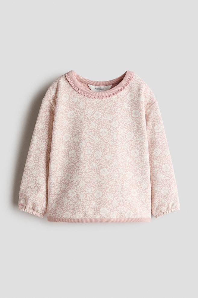 Une vue de face montre un pull en coton rose orné dâun imprimé floral blanc sur lâensemble, avec des épaules tombantes et des manches longues terminées par des poignets étroits élastiqués. Le vêtement présente un col rond côtelé agrémenté dâune délicate bordure façon dentelle, ainsi quâun ourlet côtelé assorti. - 1