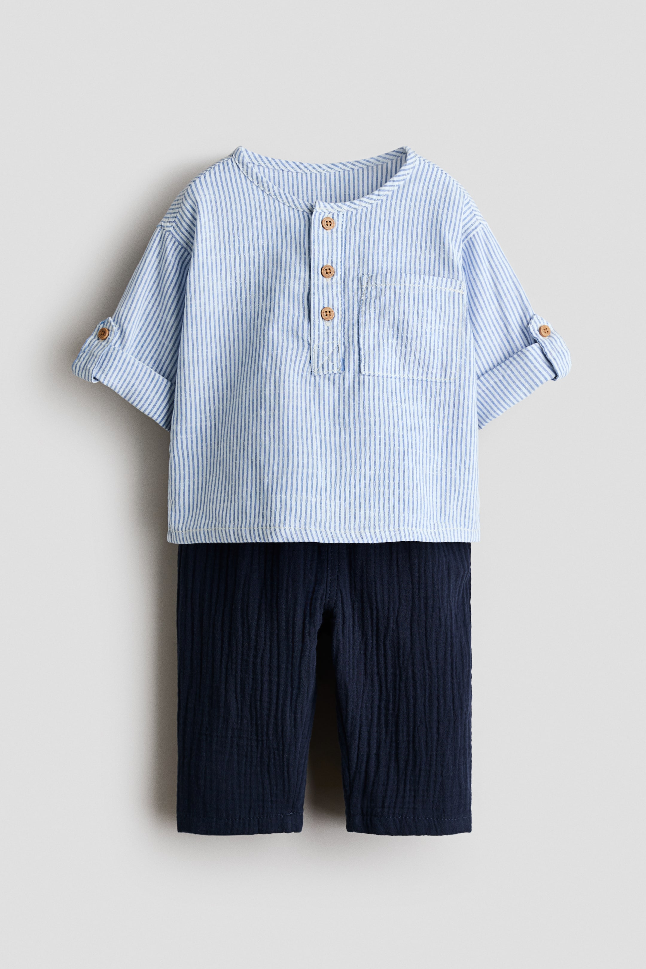 View larger image: A baby's two-piece outfit is showcased at the front, featuring a light blue and white striped shirt with a band collar, a three-button placket, a chest pocket, and rolled-up sleeves secured with tabs. Dark navy blue textured pants complete the set.