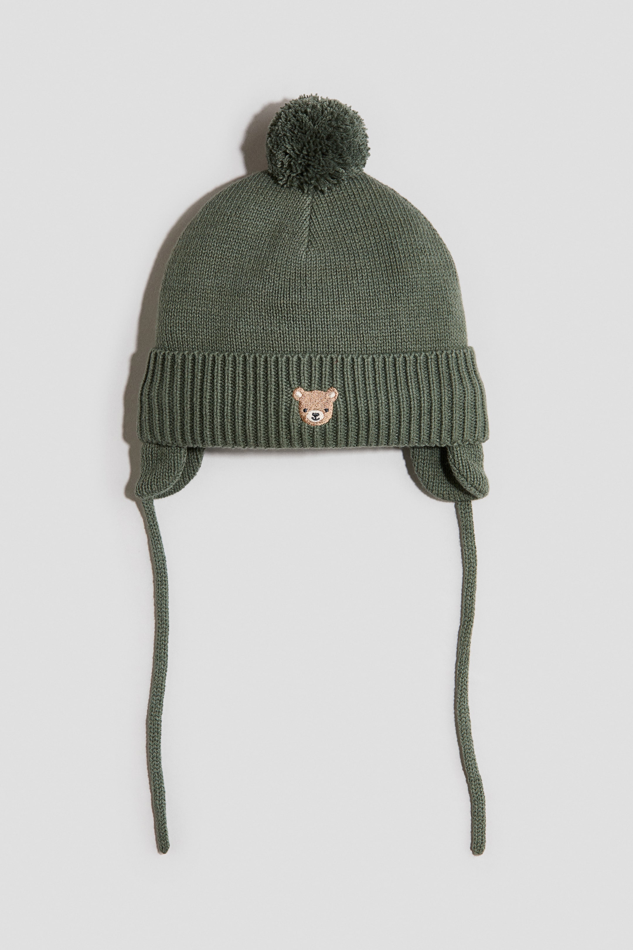 Visualizza l'immagine ingrandita: Un cappello a maglia verde scuro è presentato frontalmente, caratterizzato da un pompon testurizzato sulla parte superiore e da una fascia a coste. Mostra paraorecchie con lacci in maglia e sul davanti della fascia a coste è visibile un piccolo volto di orso ricamato.