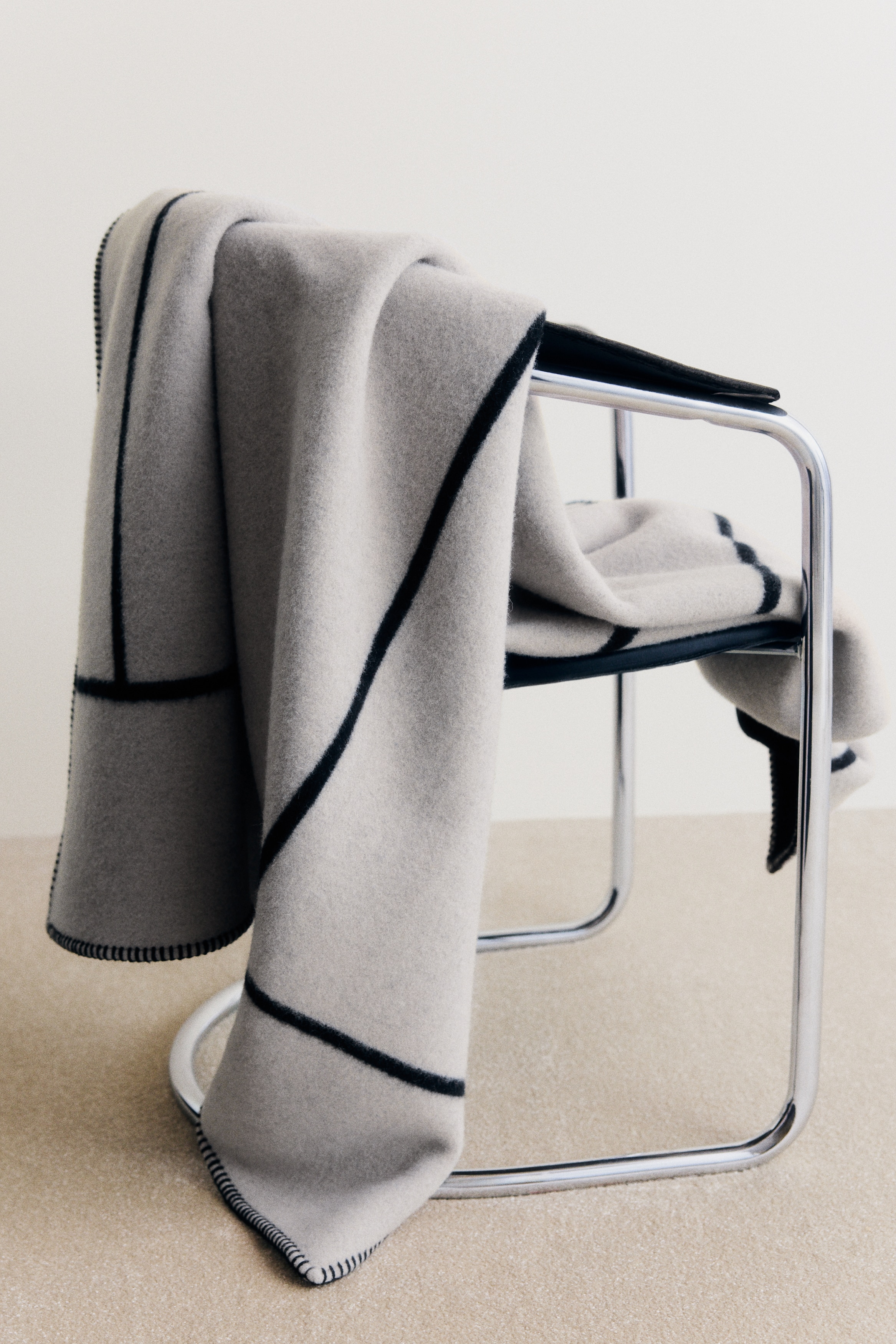Une couverture épaisse se présente à l'avant, arborant un tissage en laine mélangée gris clair avec un motif croisé en jacquard noir. Ses bords sont finis avec un point de couverture noir.