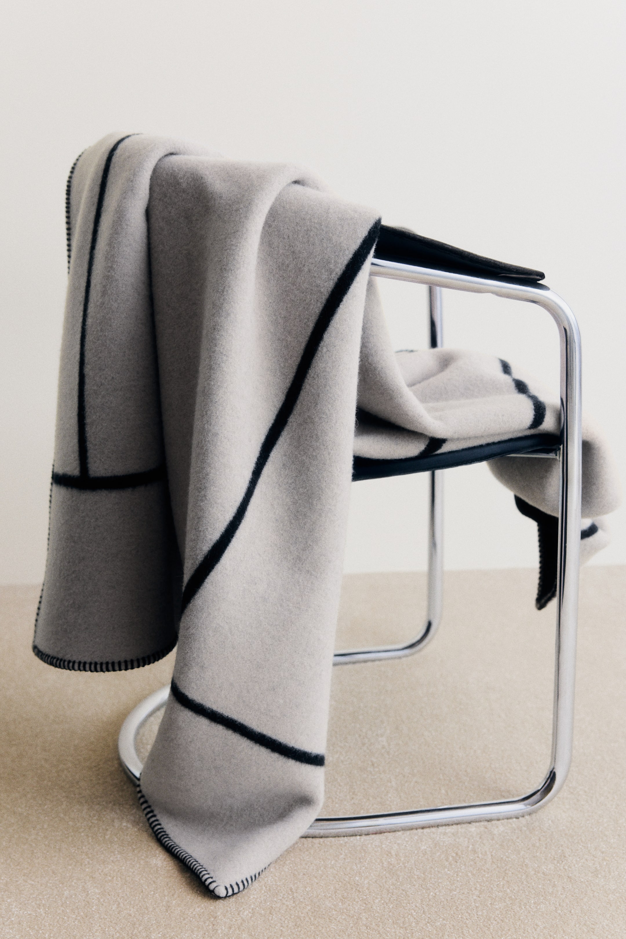 Ver imagen más grande: Una manta gruesa de mezcla de lana en gris claro cae sobre una silla moderna con estructura cromada, mostrando un atrevido patrón jacquard geométrico en negro. Los bordes de la manta están rematados con un pespunte decorativo oscuro en contraste.