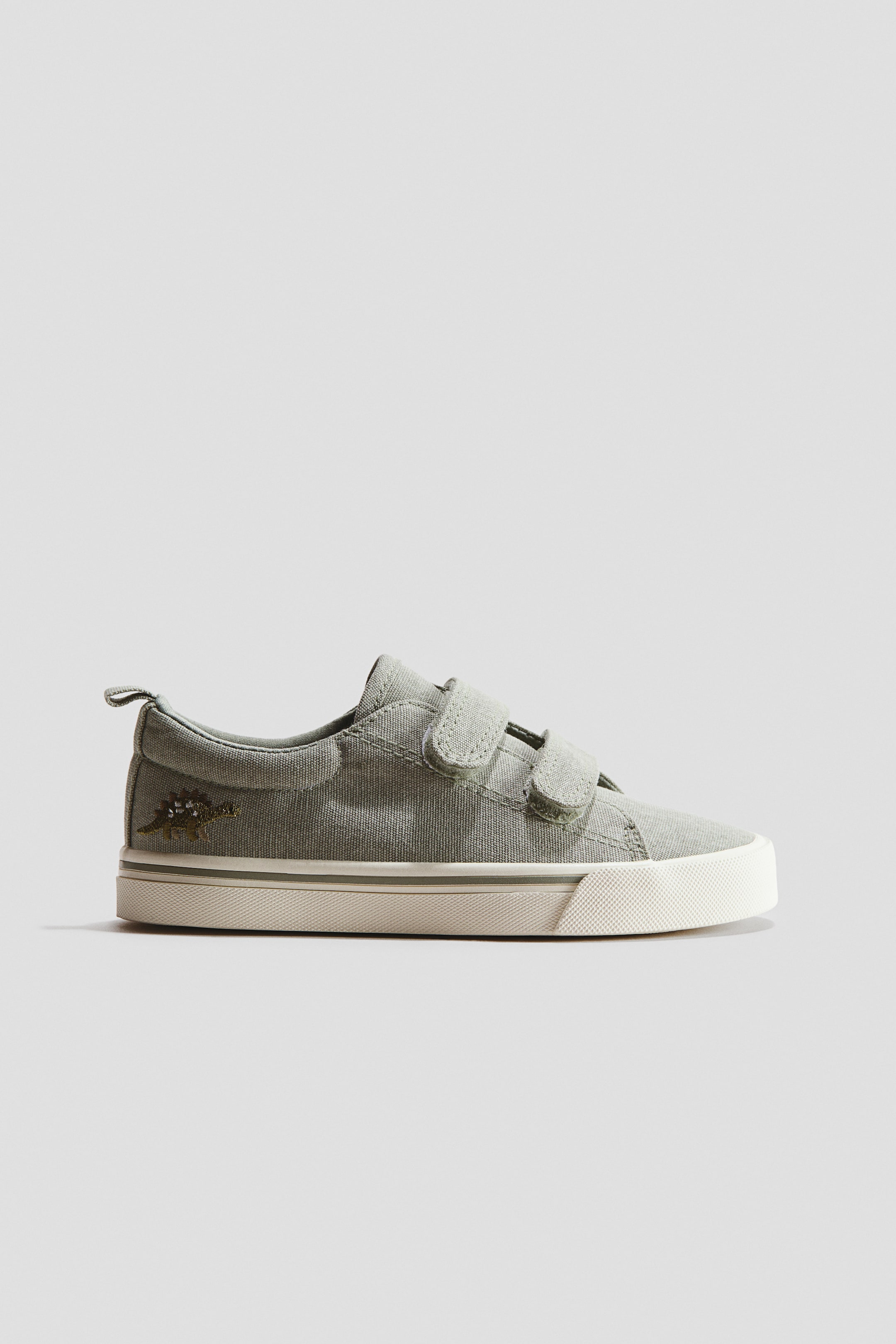 Agrandir l'image: Des baskets basses en toile de coton vert sauge sont présentées de côté, légèrement inclinées vers l'avant, avec deux languettes à scratch à l'avant, une boucle à l'arrière, un motif de dinosaure brodé sur le côté, et une semelle en caoutchouc texturé blanc.