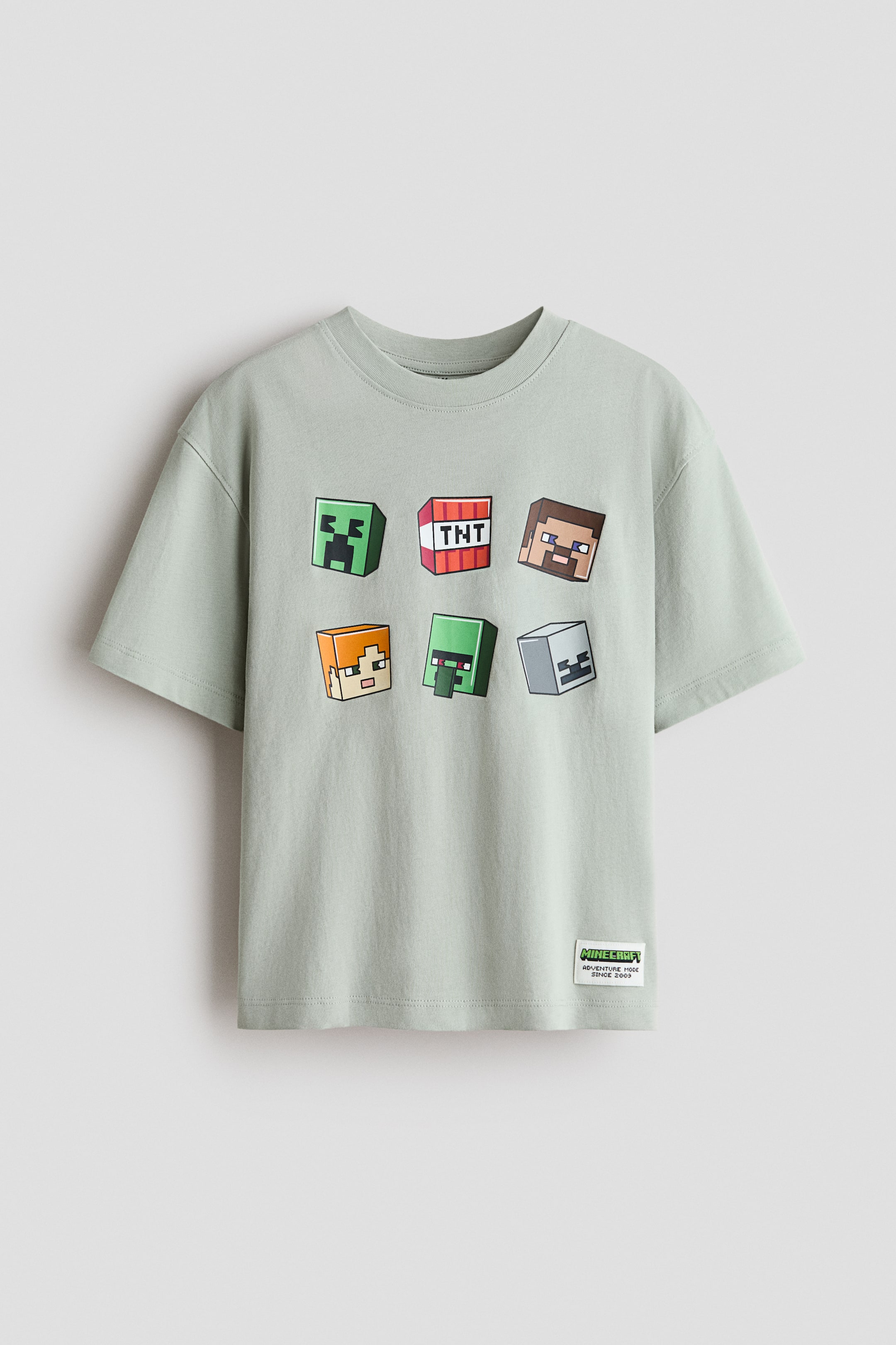 View larger image: A light green t-shirt featuring a ribbed crew neck and short sleeves, with its front displaying a graphic print of six square, block-like characters and items. A small woven label with "Minecraft" text is attached to the bottom left hem.