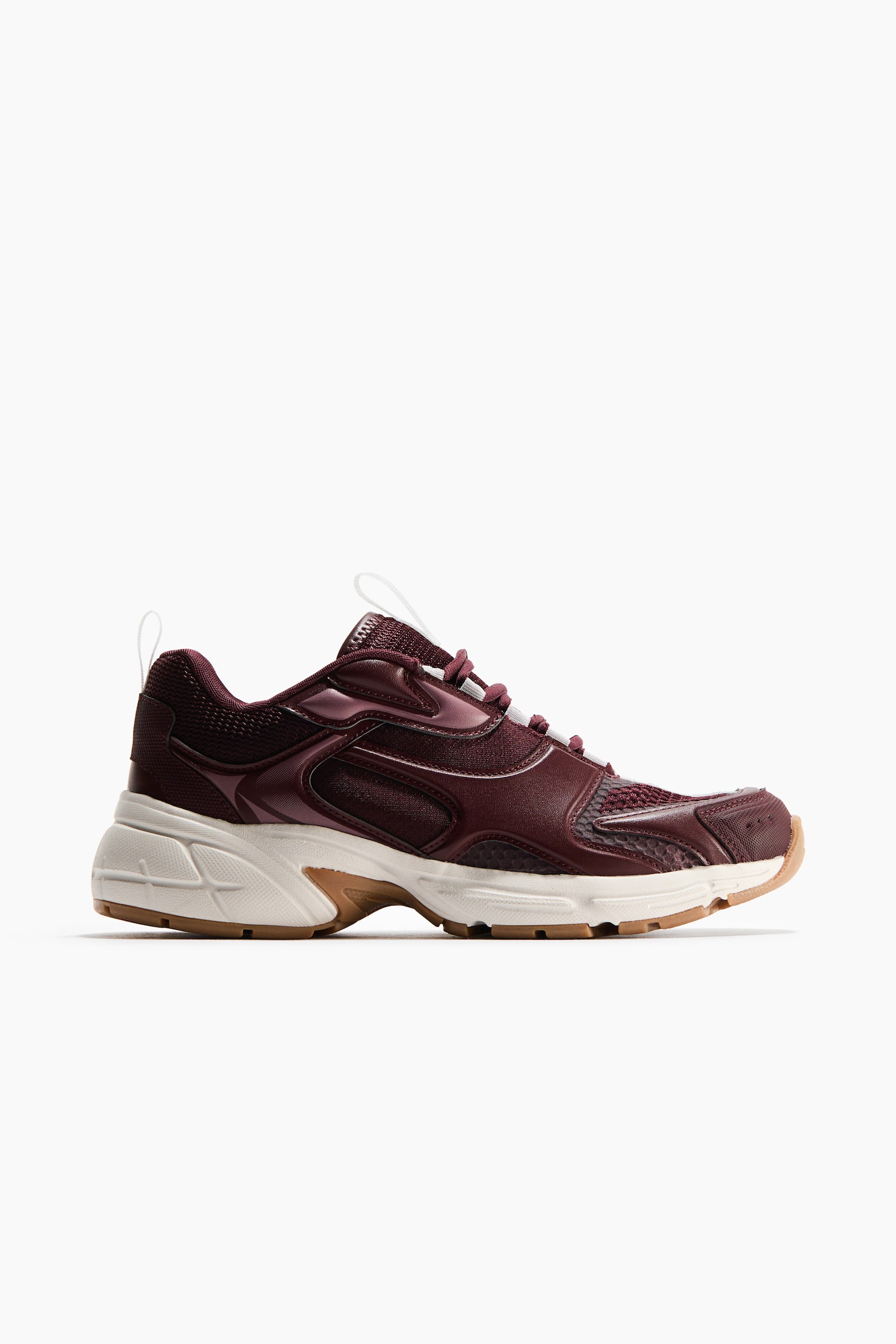 נעל סניקרס בצבע בורדו מציגה את הפרופיל הצדדי שלה, עם פאנלים מרושתים במרקם וטקסטורות סינתטיות מבריקות. היא מעוצבת עם סוליה עבה בצבע לבן-שבור, סוליה תחתונה מגומי בצבע שזוף ושרוכים לבנים בחלק העליון. - 1
