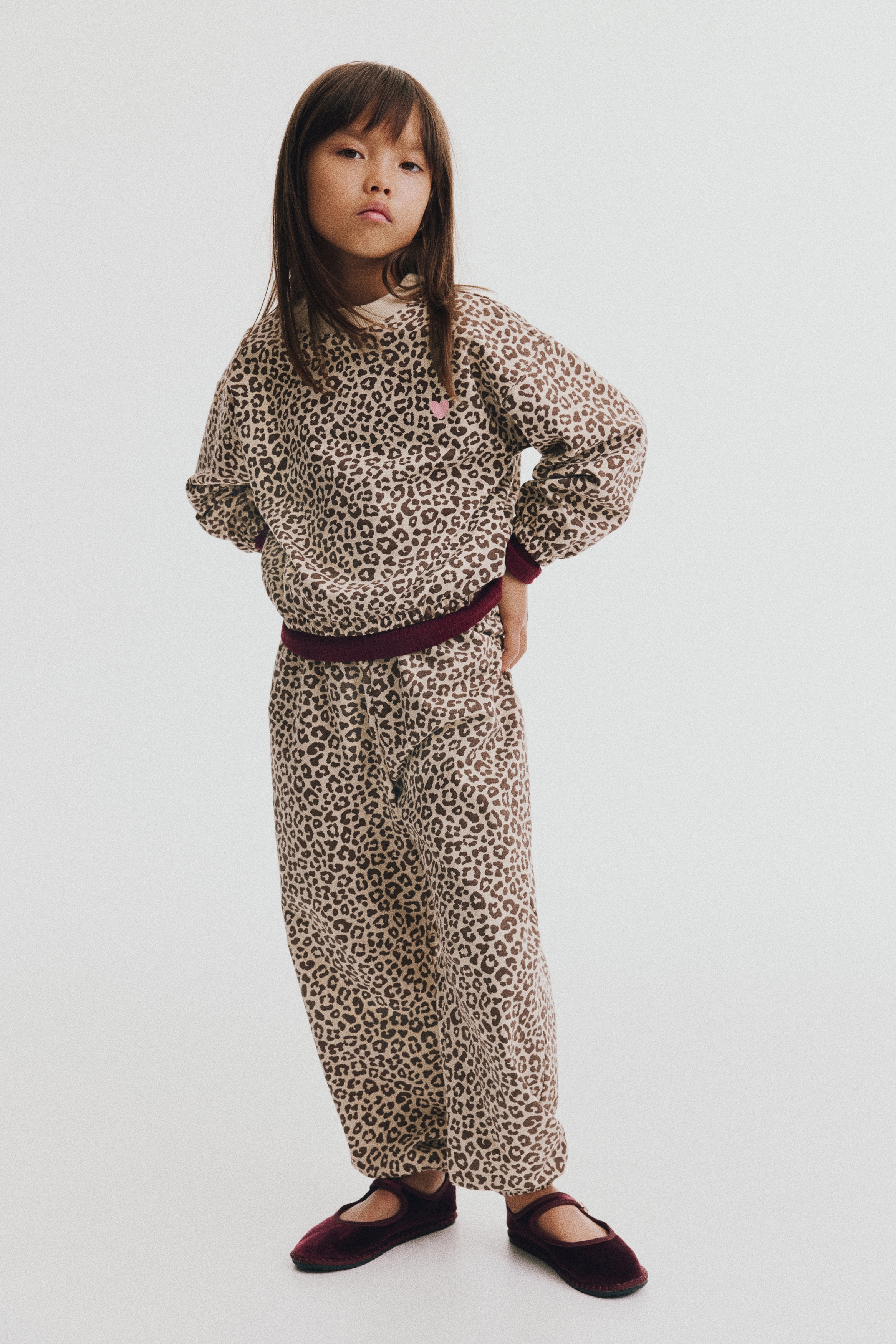 A front-facing view shows a matching leopard-print sweater and track pants set. The sweater features a rib-trimmed round neckline, dropped shoulders, voluminous sleeves with elasticated cuffs, and a small pink heart on the chest, while the pants feature an elasticated waist and gathered hems.