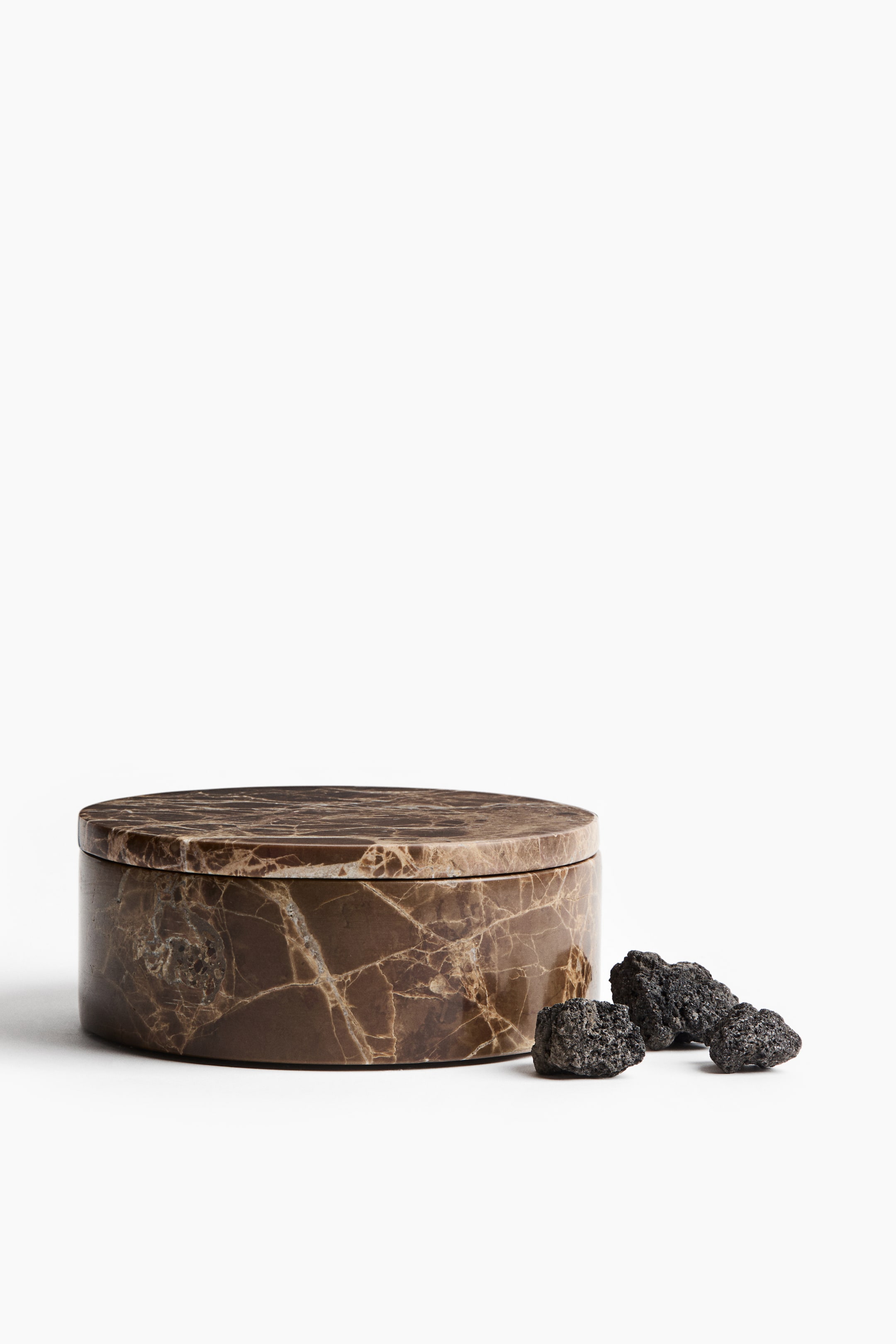 View larger image: A round, low-profile container made of brown marble with prominent white and tan veining is shown with a matching flat lid from a front-facing perspective. Three small, porous black lava stones are resting on the surface directly next to the container.