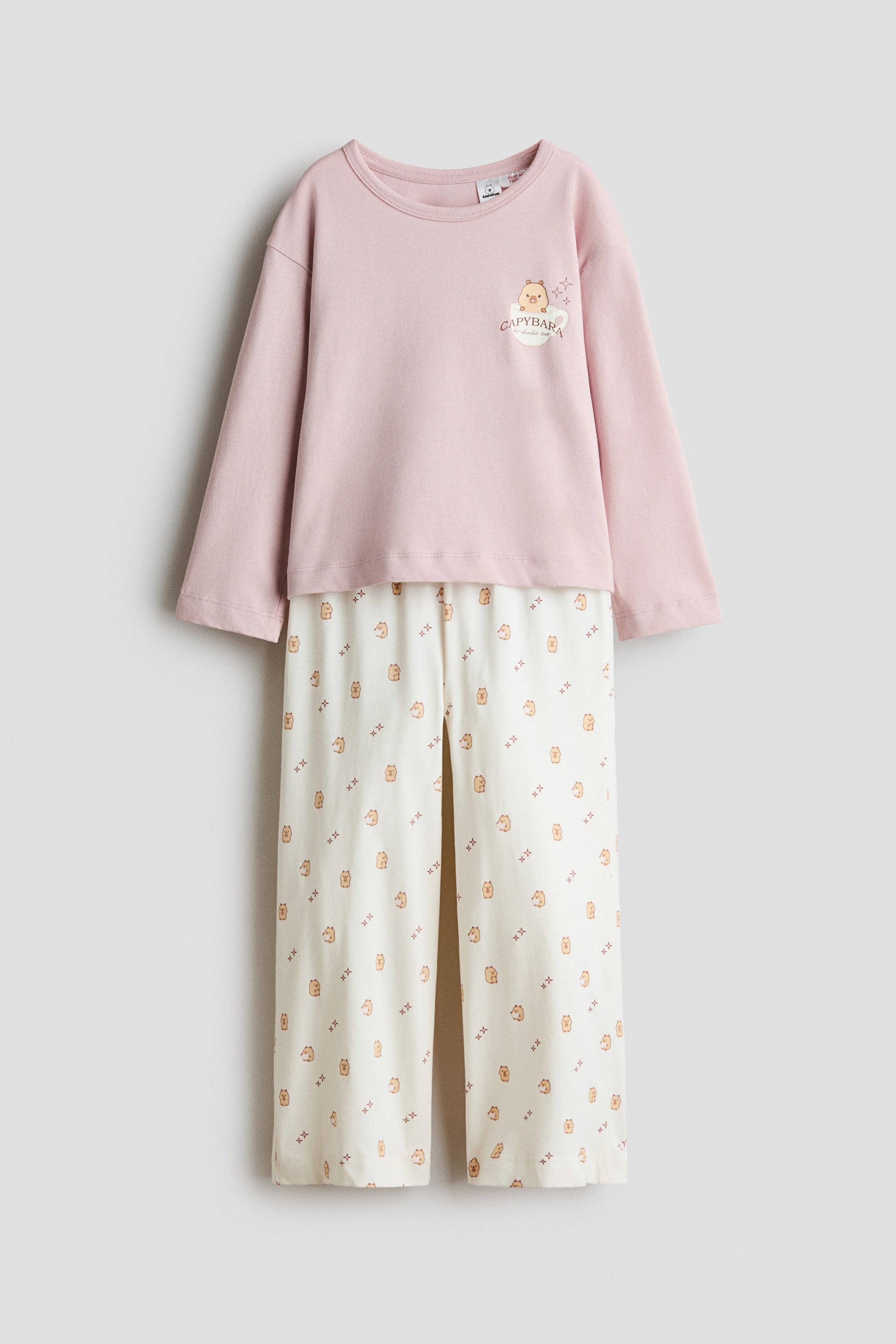 Größeres Bild anzeigen: Ein zweiteiliges Pyjama-Set wird in der Vorderansicht gezeigt und besteht aus einem hellrosa Langarmshirt mit Rundhalsausschnitt und einem kleinen Capybara-Motiv auf der linken Brust. Die dazu passende cremefarbene Hose zeigt ein Allover-Print aus kleinen Capybaras und kreuzförmigen Motiven.