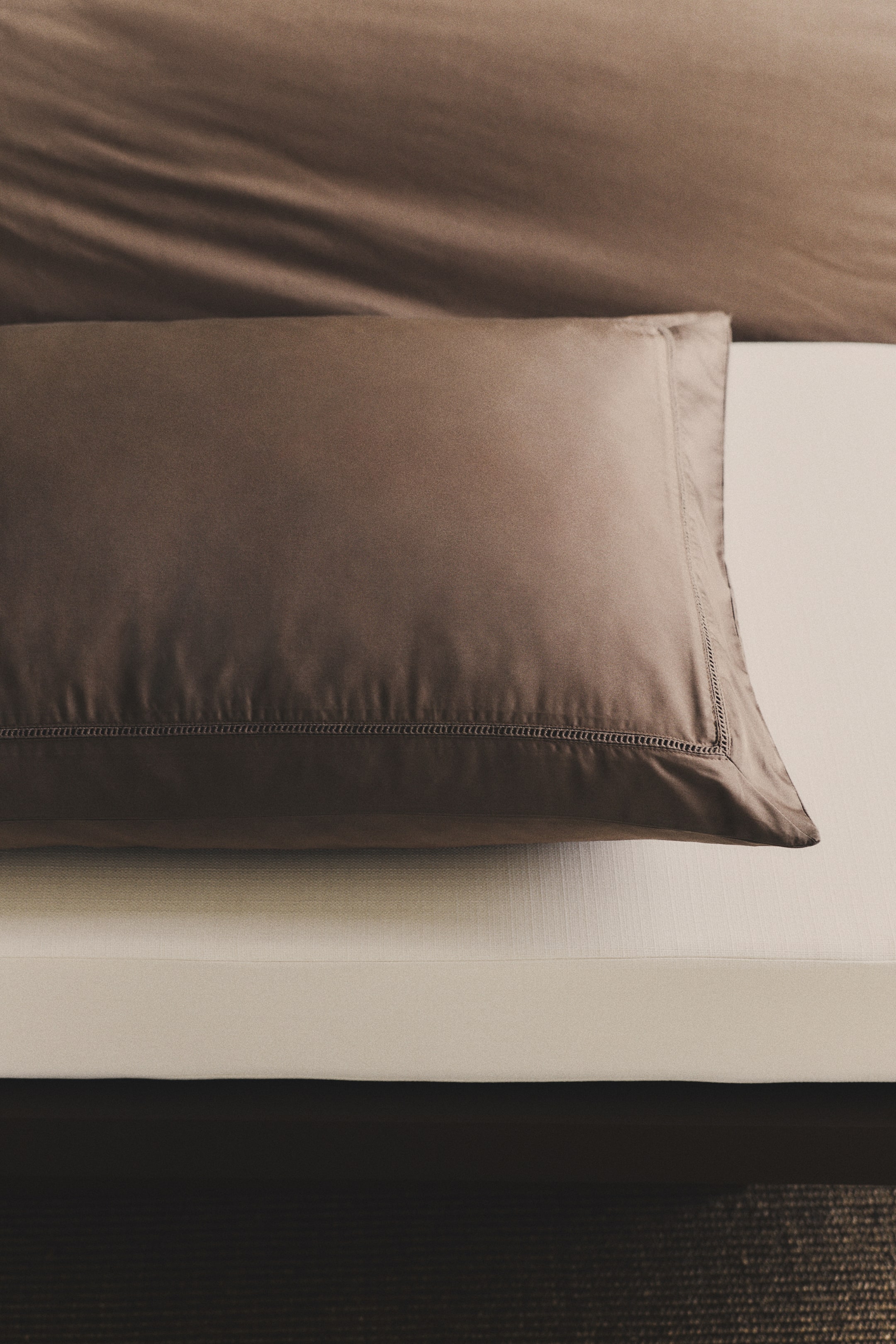Agrandir l'image: Une taie d'oreiller marron présente son devant, mettant en valeur un tissu en satin de coton à fils fins et lisses. Des bordures en dentelle échelle ton sur ton ornent ses bords, et elle dispose d'une ouverture à rabat.