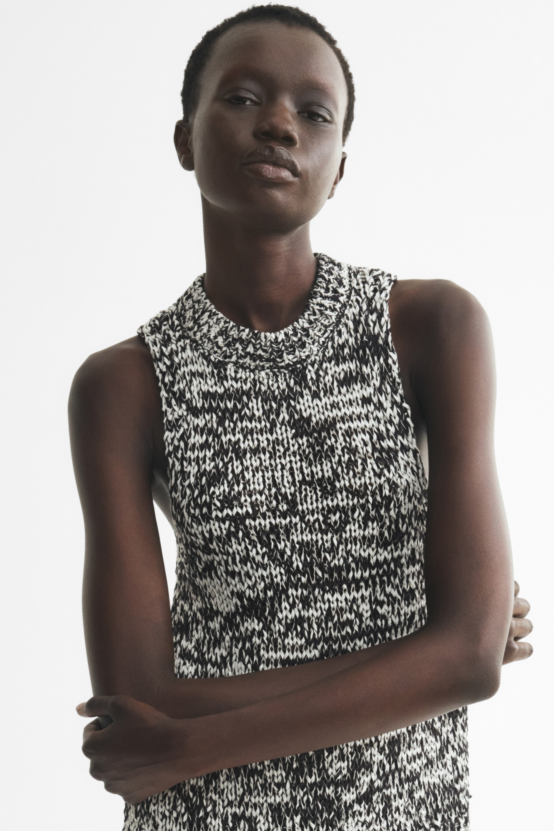 Agrandir l'image: Un débardeur en maille texturée à motif chiné noir et blanc, avec un col rond côtelé et de larges emmanchures, porté par le mannequin. Le mannequin est debout, les bras croisés.