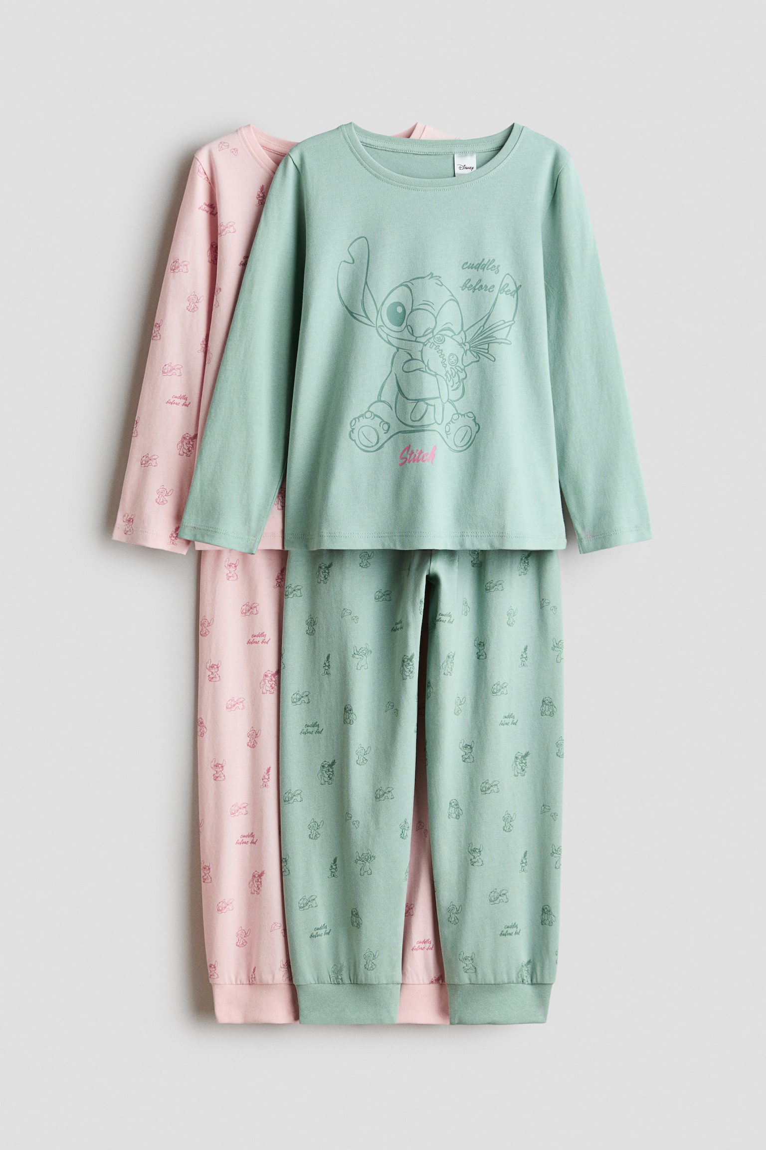 Zwei Pyjama-Sets aus Baumwoll-Jersey hängen in frontaler Ansicht, wobei ein hellgrünes Set über einem hellrosa Set liegt. Das hellgrüne Set zeigt ein Langarm-Oberteil mit Rundhalsausschnitt, grossem Stitch-Print mittig vorne sowie die dazu passenden Hosen mit Allover-Print und Rippbündchen. Das dahinter liegende hellrosa Set zeigt einen passenden Allover-Charakterprint sowohl auf dem Langarm-Oberteil als auch auf der Hose.