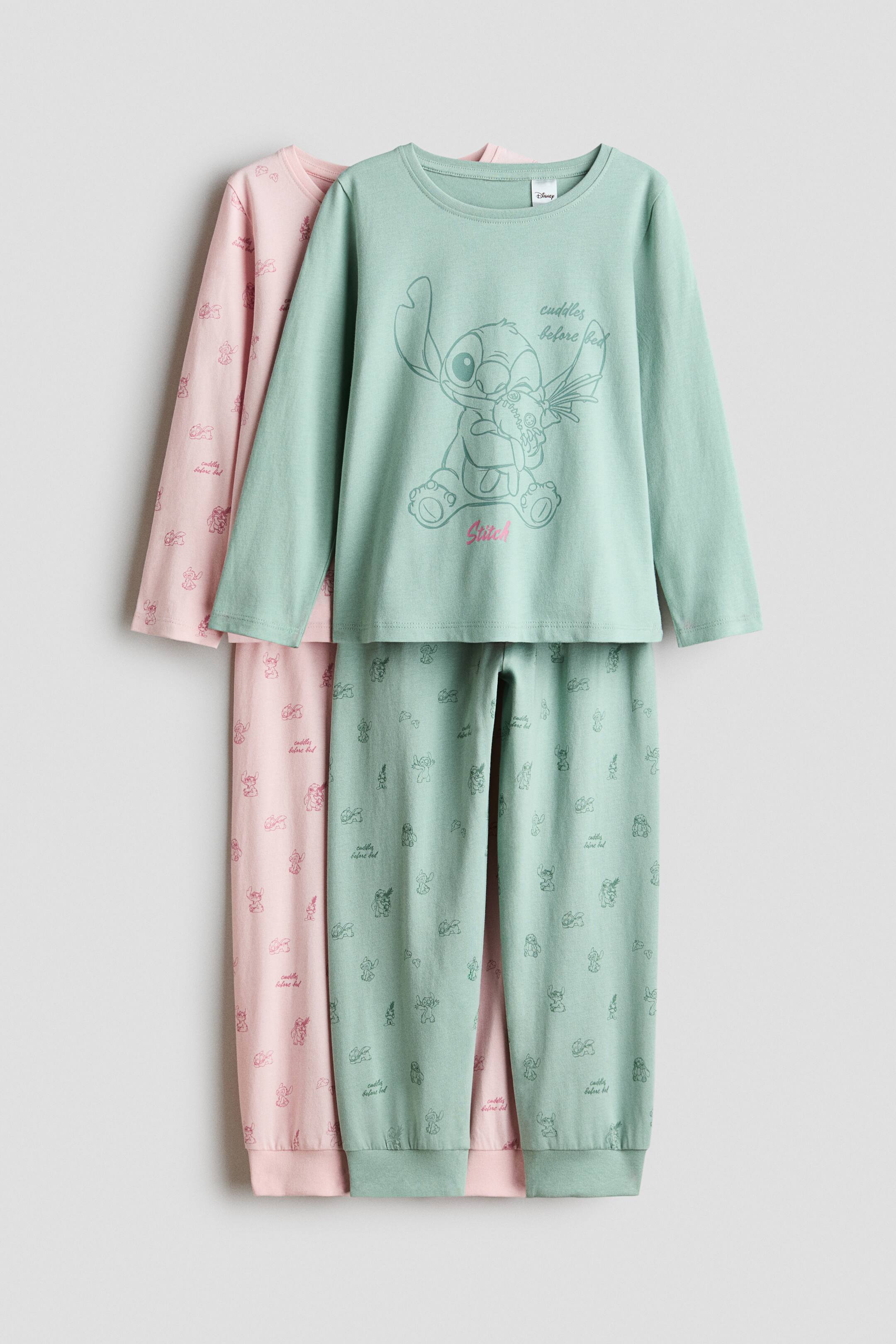 View larger image: Two cotton-jersey pyjama sets are hanging in a front-facing view, with a light green set layered over a light pink set. The light green set features a round-neck, long-sleeve top with a large central Stitch graphic and matching pants with an all-over print and ribbed cuffs. The light pink set layered behind shows a matching all-over character print on both the long-sleeve top and pants.