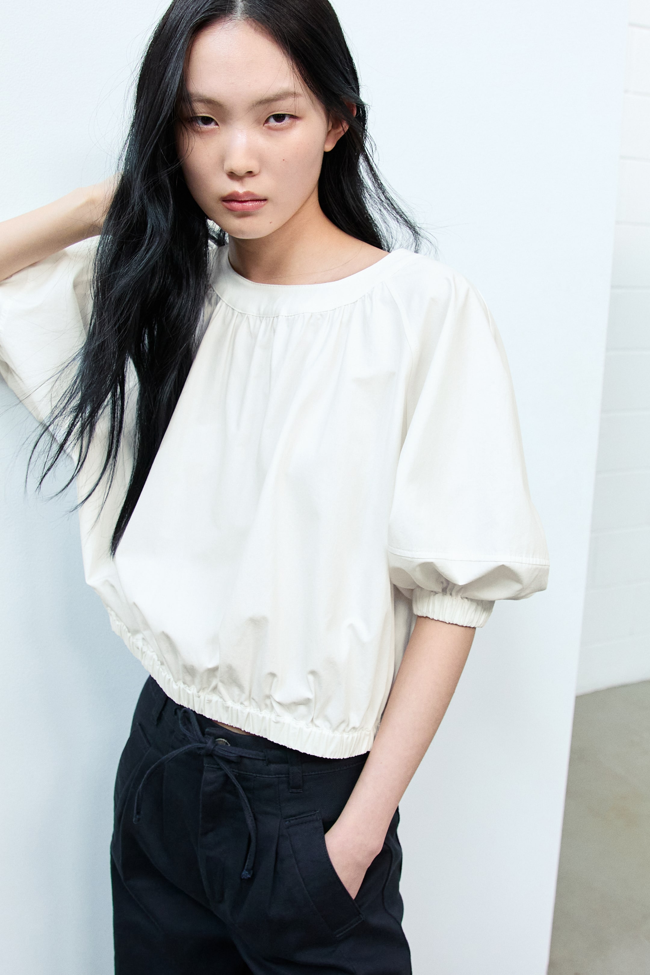 View larger image: An off-white cropped top featuring a gathered round neckline and voluminous raglan sleeves with elasticated cuffs. Its elasticated hem rests just above the waistband of black pants.