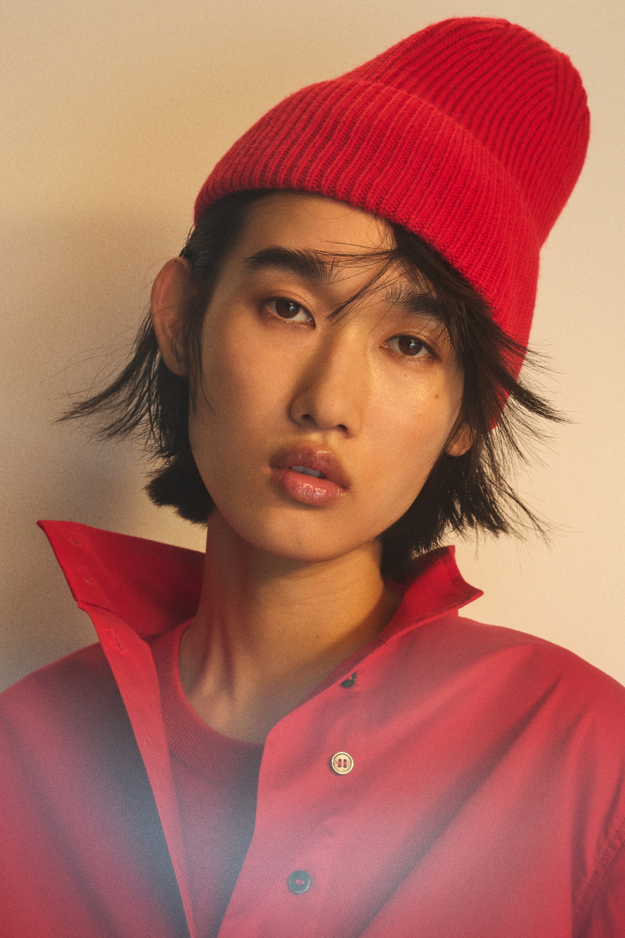 View larger image: A bright red beanie in a soft rib-knit with a folded turn-up hem is being worn by the model. The beanie is positioned high on the head and coordinates with a red collared shirt.