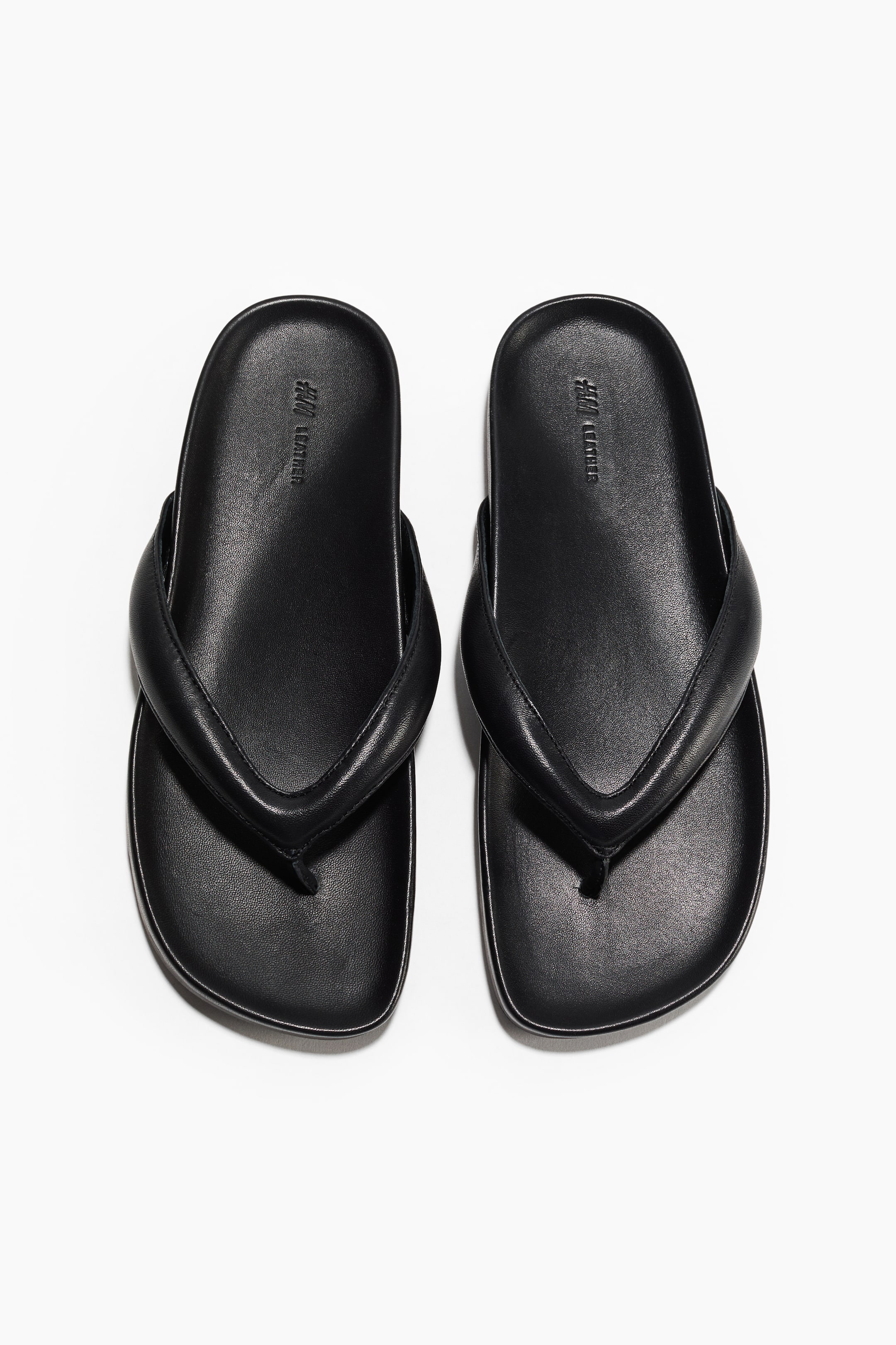 View larger image: A pair of black leather flip-flops is shown from the front, highlighting angled toes, cushioned straps, and a slim toe post. The footbeds are embossed with "H&M LEATHER."