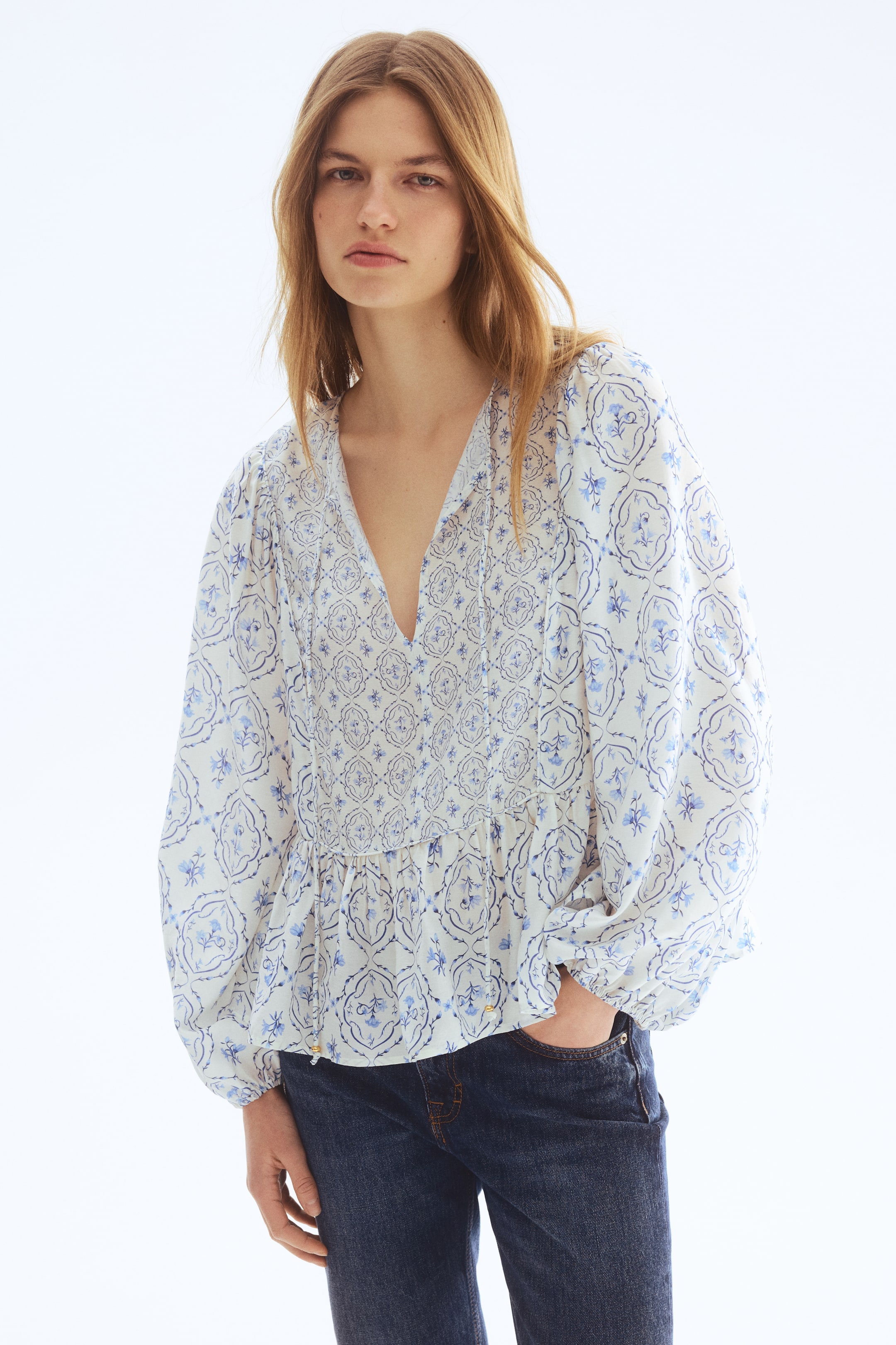 Grotere afbeelding bekijken: Een witte blouse met blauwe bloemen- en geometrische patronen, een V-hals met strikjes, een geplooid voorpand boven een geplooide peplumstijl zoom en lange pofmouwen met elastische manchetten.