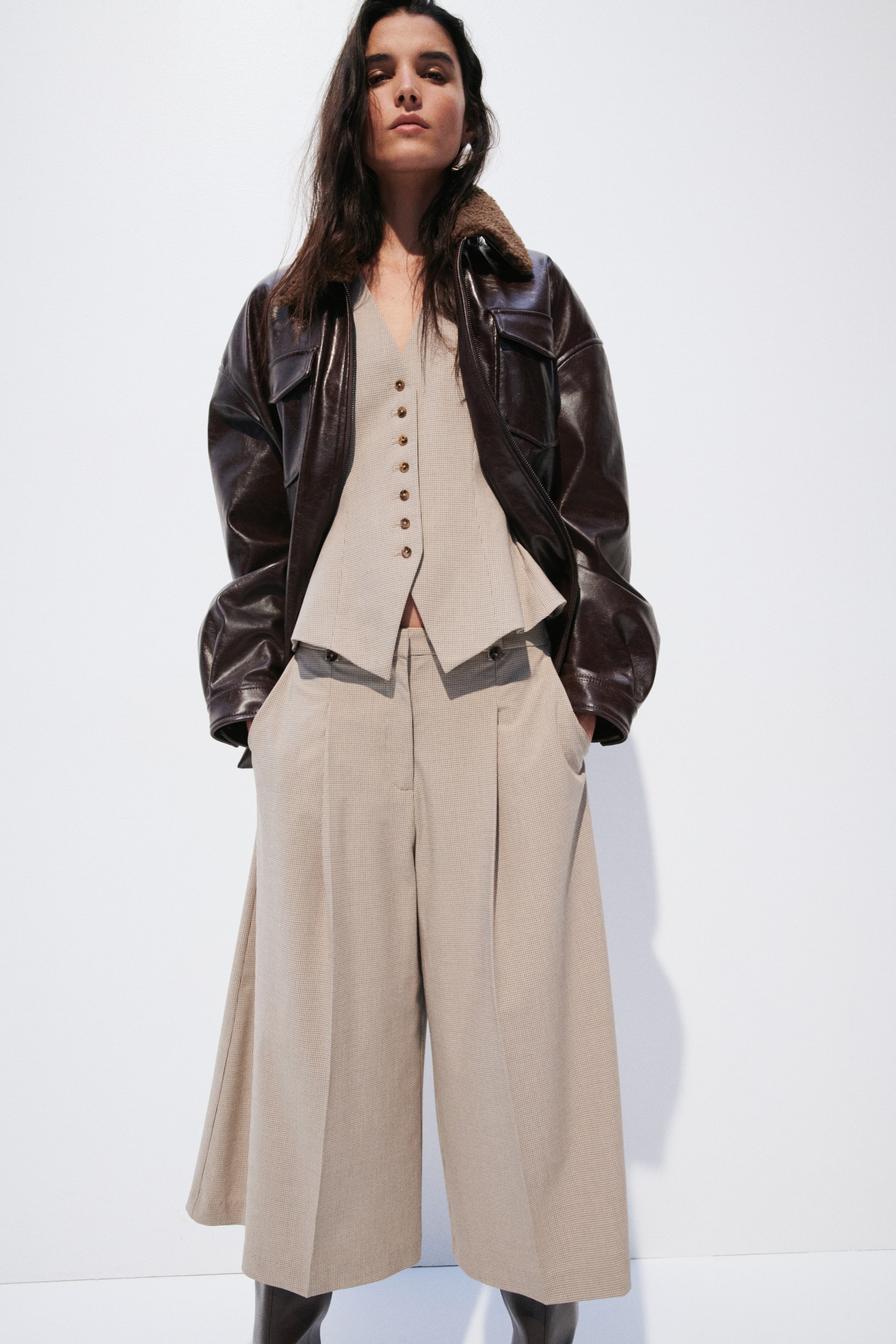 View larger image: A brown leather jacket, featuring a shearling collar and chest pockets, is worn open over a light beige V-neck waistcoat with a buttoned front and peplum hem. The waistcoat is styled with matching pleated wide-leg culottes.