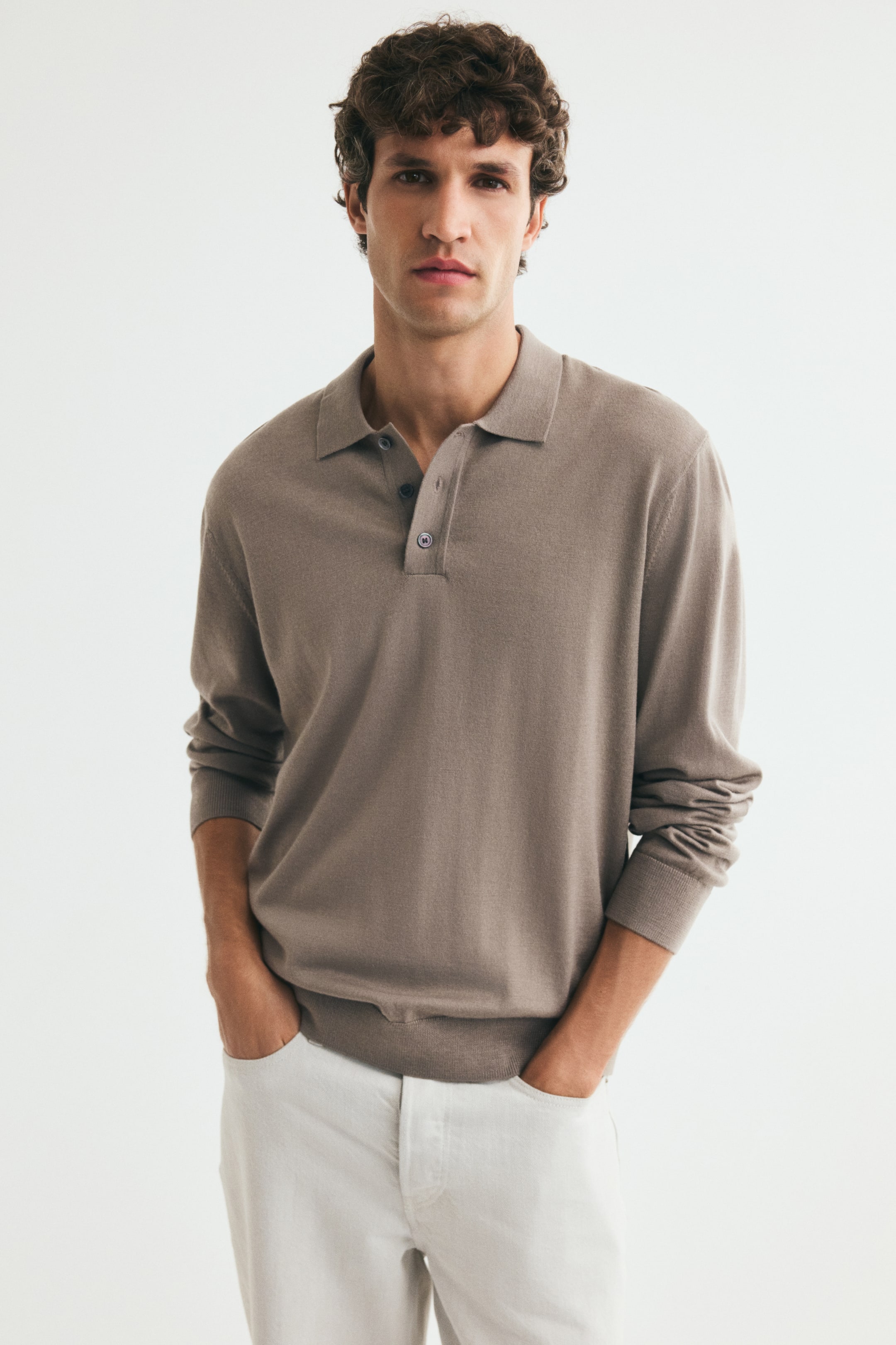 View larger image: The model is wearing a long-sleeved polo shirt in taupe fine-knit wool, with their hands in the pockets of light-coloured trousers. The shirt features a cutaway collar, a partially unbuttoned placket, and fine ribbing at the cuffs and hem.
