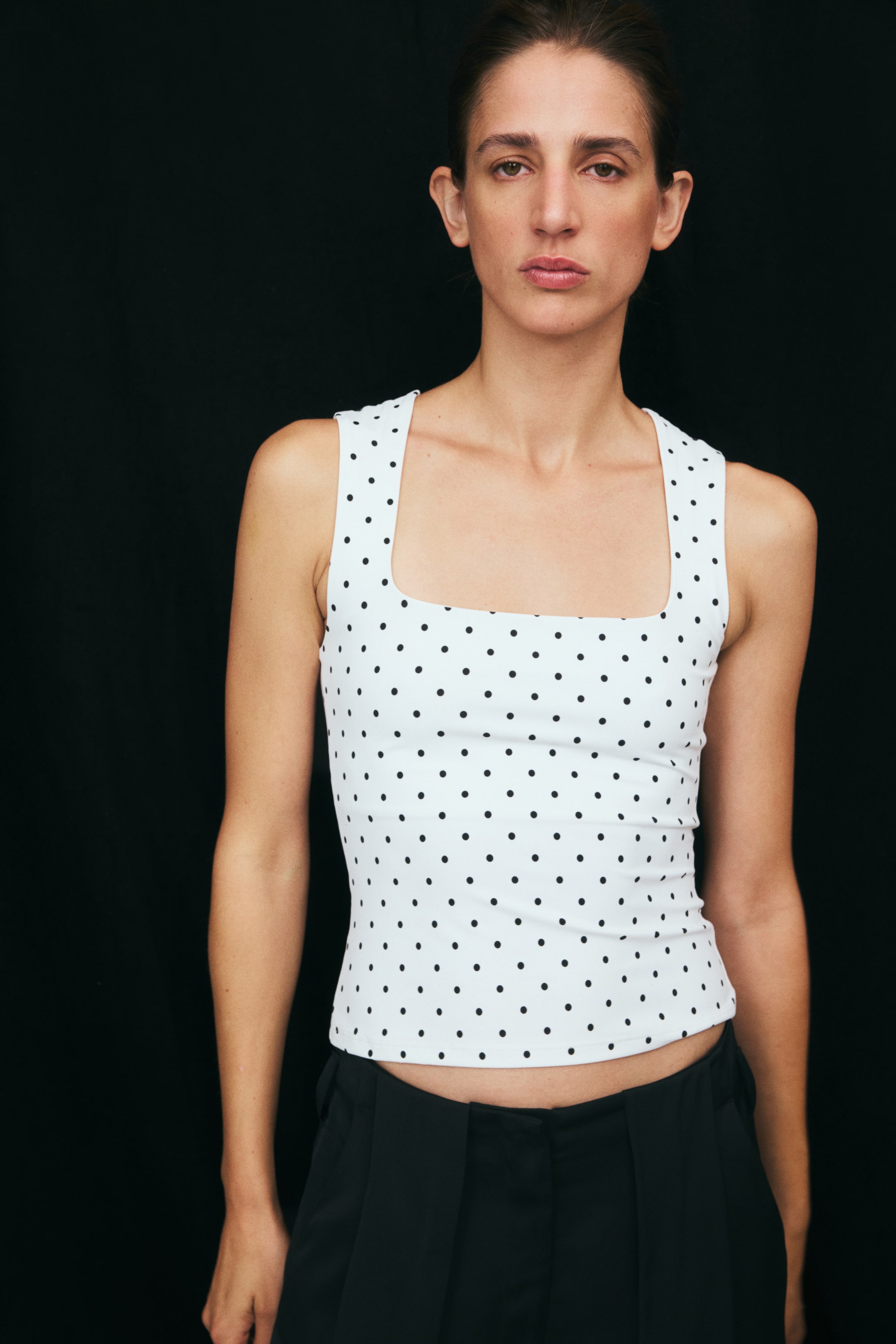 View larger image: The model is wearing a white fitted tank top with a black polka dot pattern. The sleeveless top features a square neckline and a straight hem, and is paired with black trousers.