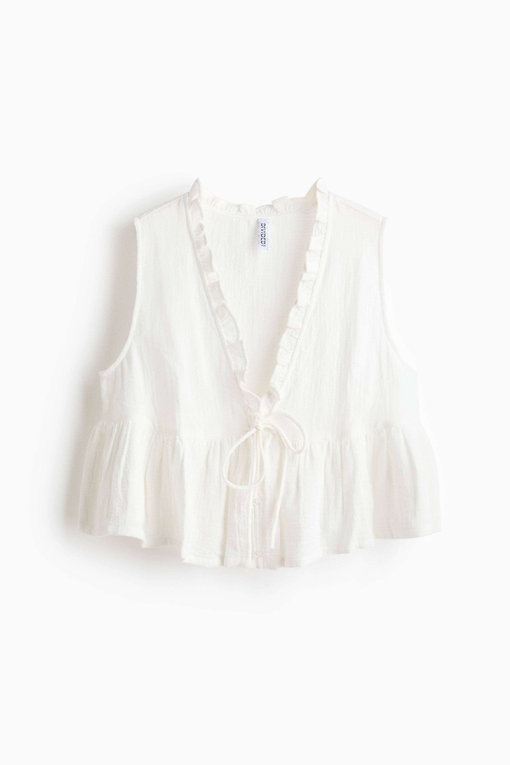 View larger image: A white sleeveless top is facing forward, featuring a ruffled V-neckline and a tie-front closure over visible buttons, while displaying a gathered peplum hem and a crinkled texture.