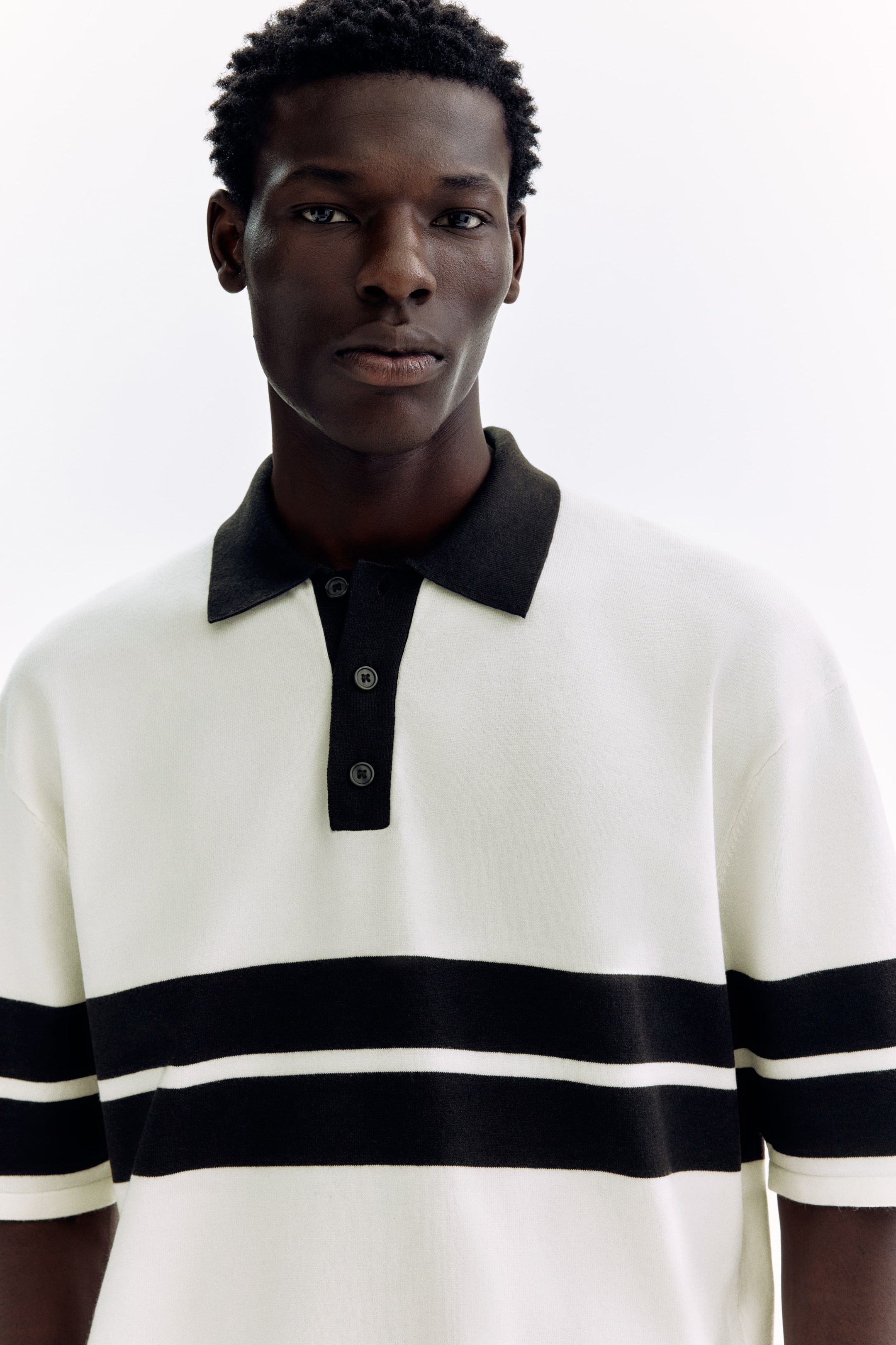 Grösseres Bild ansehen: Ein kurzärmliges Strick-Poloshirt mit einem weißen Körper, einem schwarzen Kragen, einer schwarzen Zwei-Knopf-Leiste und zwei schwarzen horizontalen Streifen über der Brust wird vom Modell getragen und präsentiert einen lockeren Sitz.