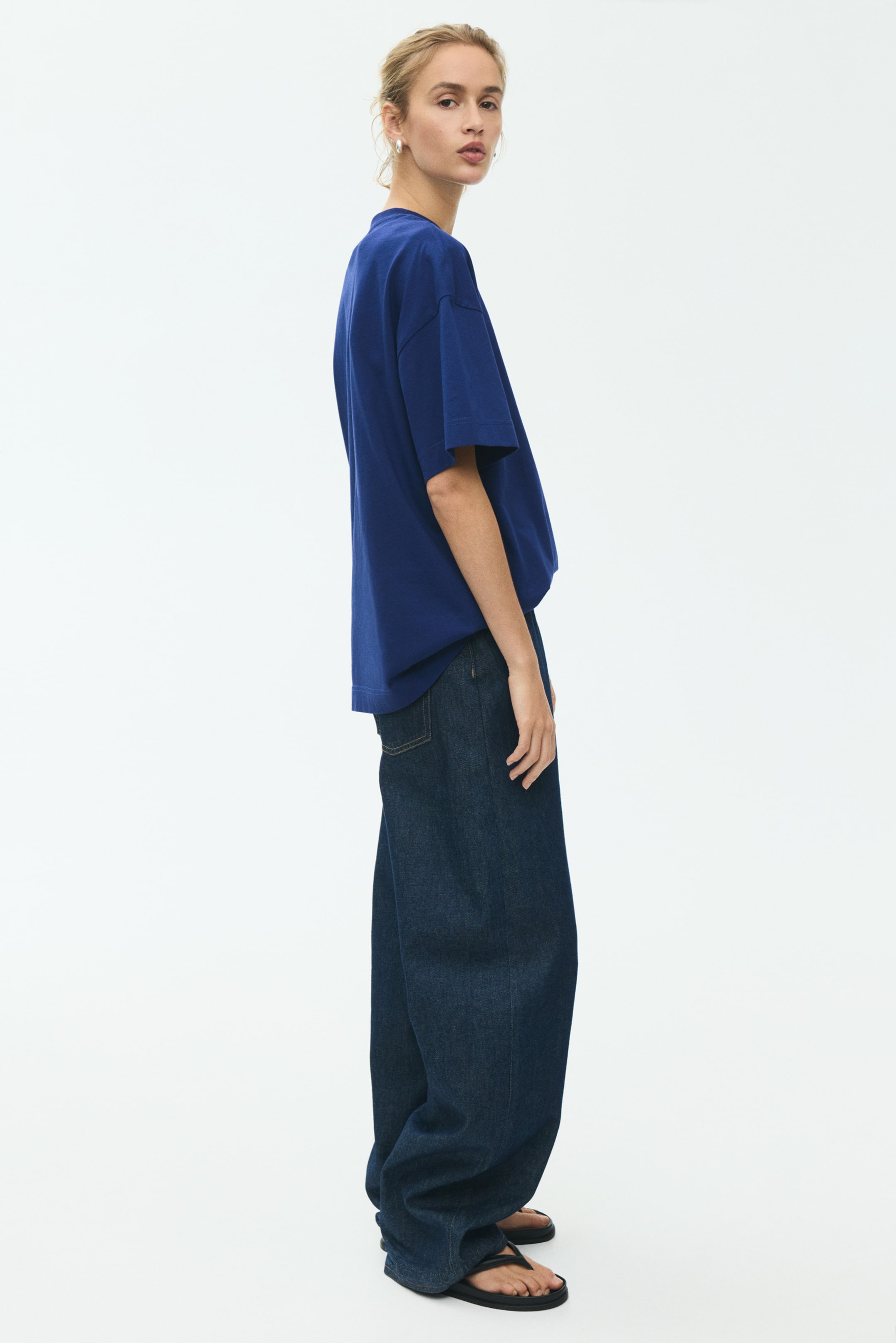 Grotere afbeelding bekijken: Een loszittend, donkerblauw T-shirt met ronde hals en wijde korte mouwen wordt gedragen, gezien vanuit een zijprofiel, met de zoom ingestopt in donkergewassen, wijde jeans. De jeans hebben een relaxte pasvorm en vallen lichtjes op de enkels.