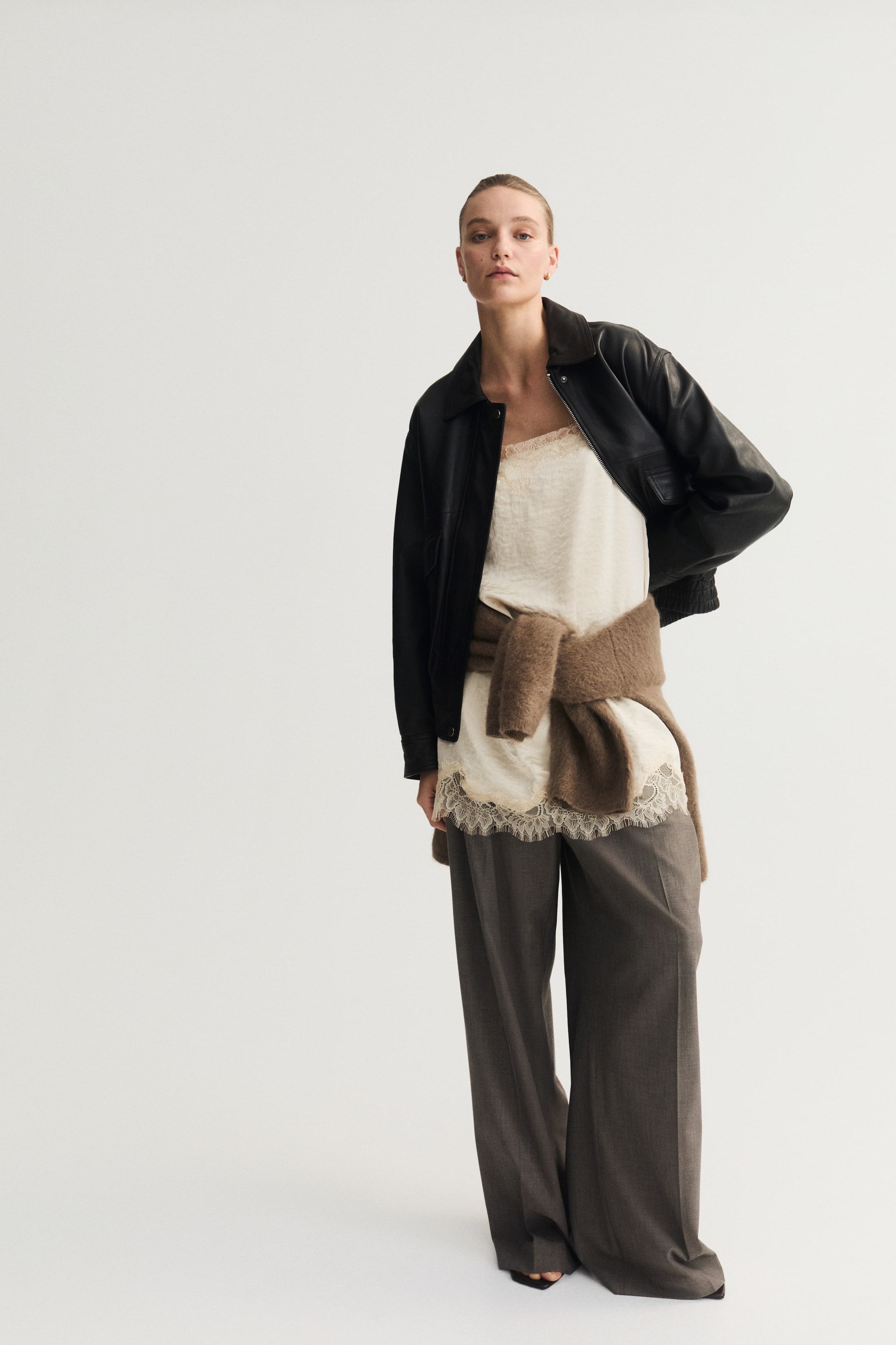 הצגת תמונה גדולה יותר: מעיל עור שחור עם צווארון קלאסי ושרוולים מצולעים נלבש פתוח מעל גופיית סליפ בצבע קרם, הכוללת תחרה בקו הצוואר ובשוליים. סוודר סרוג בצבע חום קשור סביב המותן, ומכנסיים רחבים בצבע אפור כהה נלבשים.