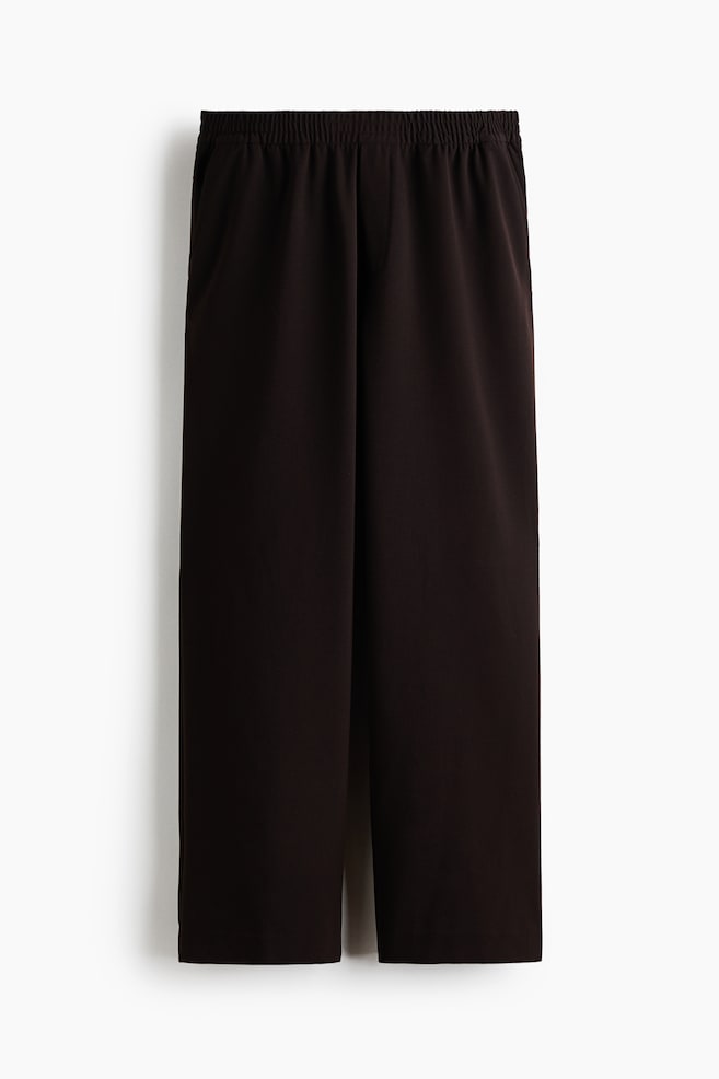 A pair of dark brown, wide-leg trousers is hanging in a front-facing view, showing a relaxed fit and a gathered, covered elastic waistband. The trousers feature a subtle mock fly and diagonal front pockets. - 1