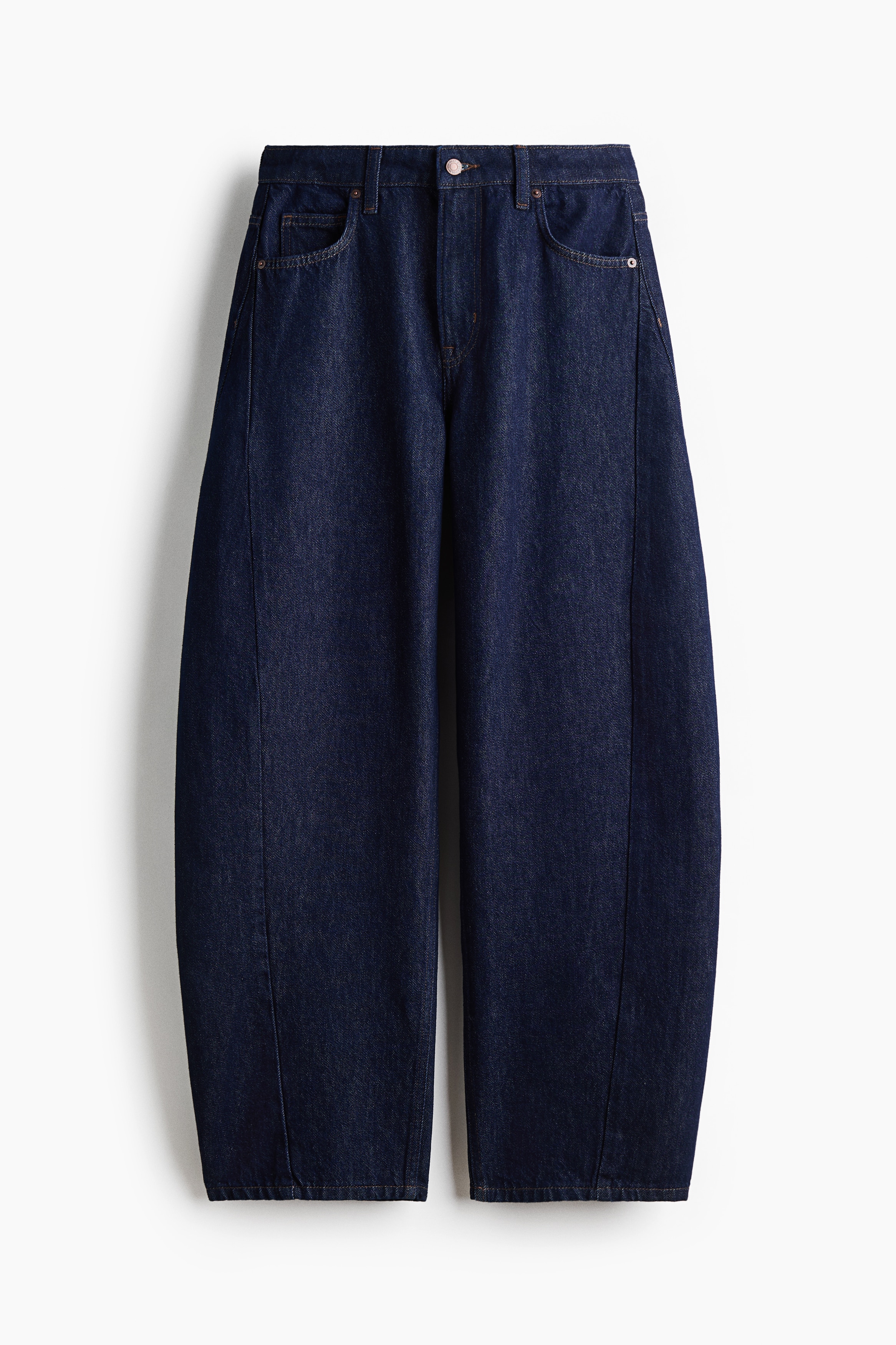 MÃ¶rkblÃ¥ jeans i denim med normal midja och bÃ¶jda, voluminÃ¶sa ben bÃ¤rs av modellen, som stÃ¥r med hÃ¤nderna i framfickorna. Jeansen stylas med en kort svart skinnjacka utan krage, en vit topp och svarta tongsandaler.