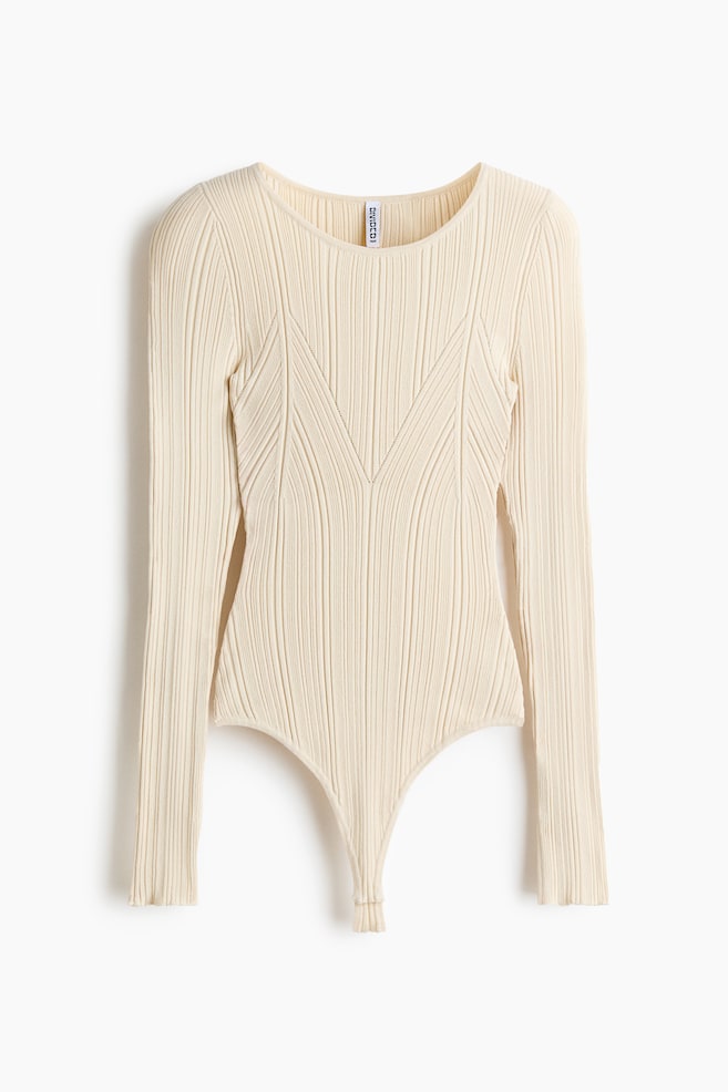 Un body a maniche lunghe color crema è mostrato frontalmente, con scollo rotondo e una texture a maglia a coste. Il design mette in evidenza un motivo decorativo a coste a V sul davanti del bustino e si completa con una silhouette da body sgambato alto. - 1
