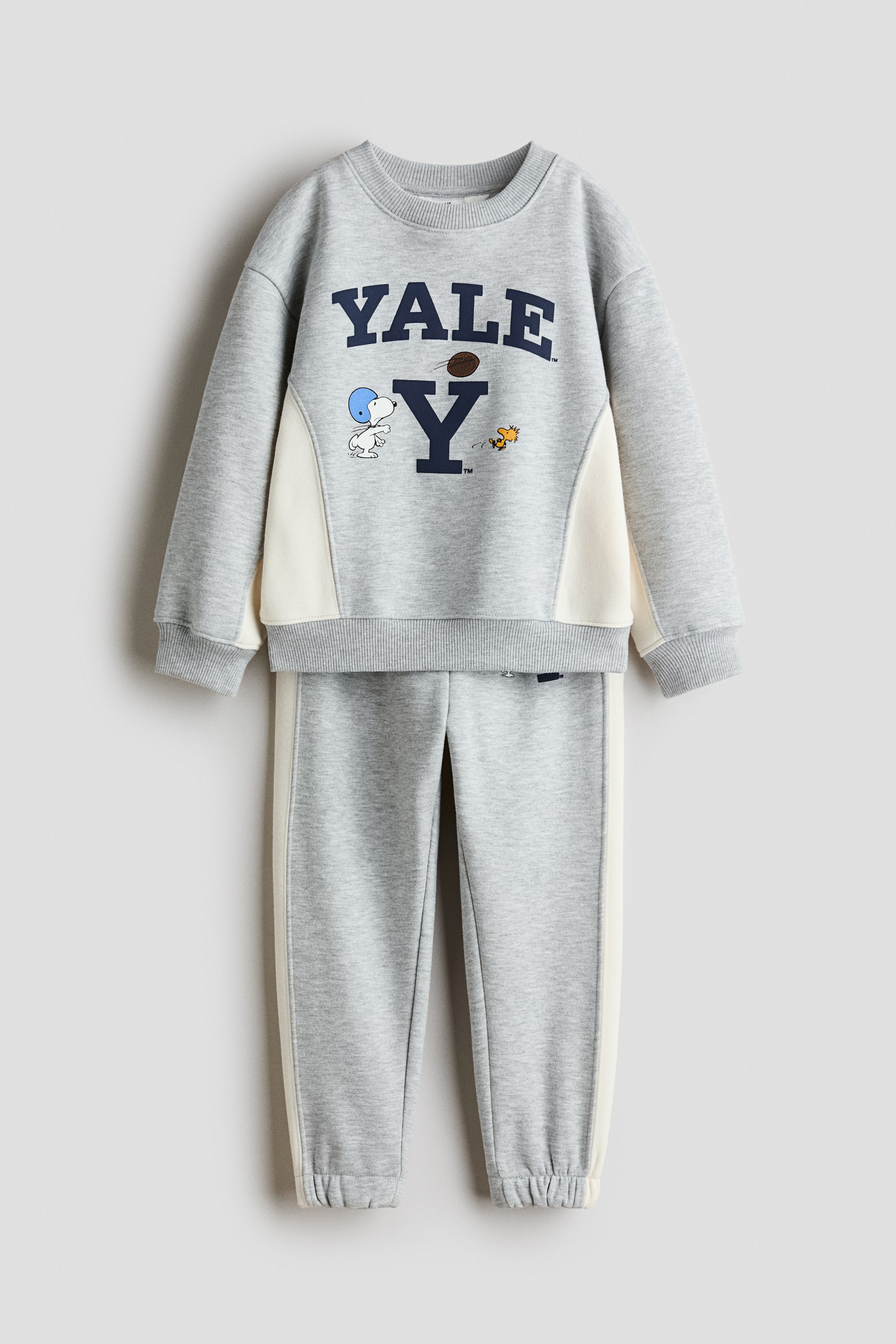 View larger image: A two-piece heather grey tracksuit set is shown from the front, featuring a crewneck sweatshirt and matching sweatpants. The sweatshirt displays a navy blue "YALE" graphic with Snoopy and Woodstock illustrations, cream-coloured side panels, and ribbed trims. The sweatpants feature matching cream-coloured side stripes and elasticized ankle cuffs.