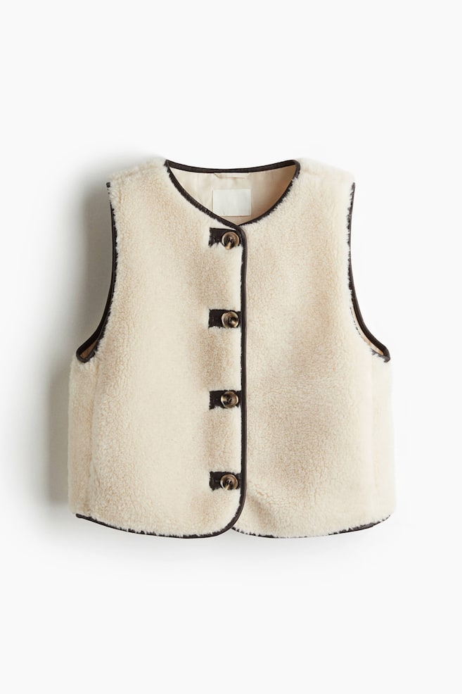 A cream-colored sherpa vest is presented from the front, showcasing dark brown piping along its rounded neckline, armholes, and curved hem. Five dark, round buttons are detailing the center front closure. - 1
