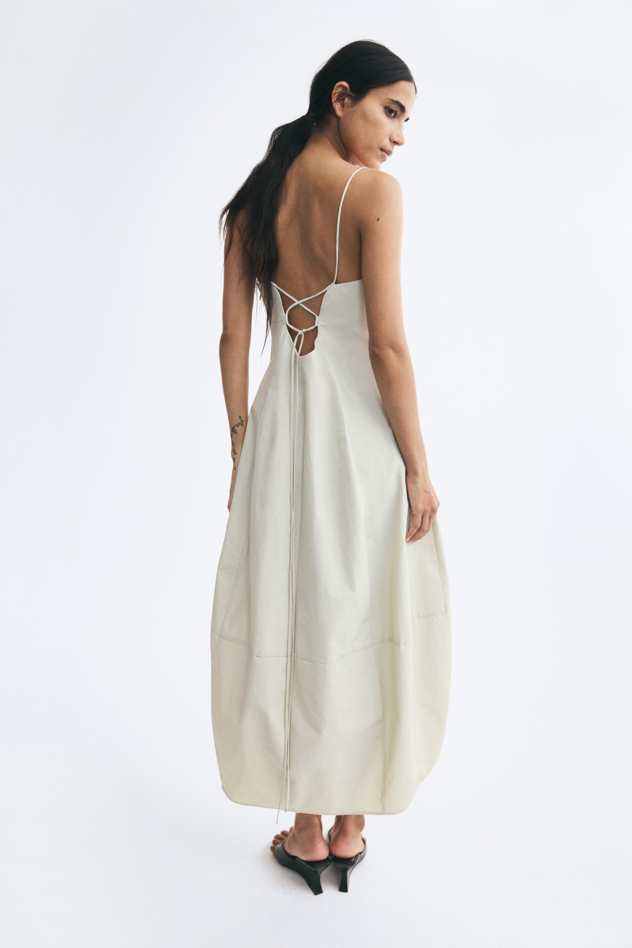 View larger image: A long, light-beige dress in a crisp fabric is worn by the model, showcasing its low-cut back with crisscrossing spaghetti straps that lace through loops and tie. The dress features a flared skirt that narrows toward the hem to create a cocoon silhouette with a prominent horizontal seam.