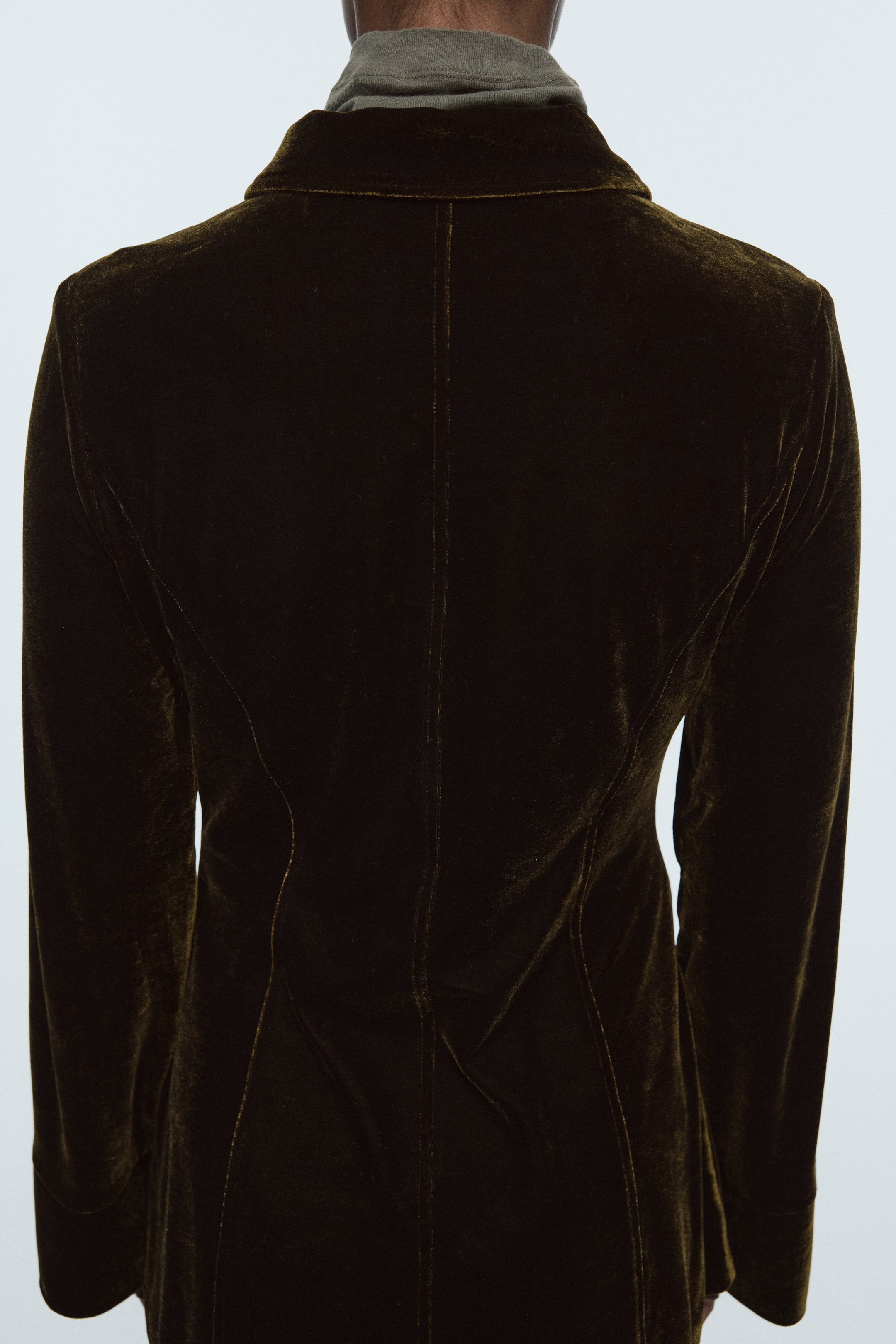 View larger image: A dark olive velvet jacket is being viewed from the back, featuring a collar, a prominent center back seam, and two curved vertical seams on either side. It is being worn over a grey mock neck shirt, visible at the neckline.