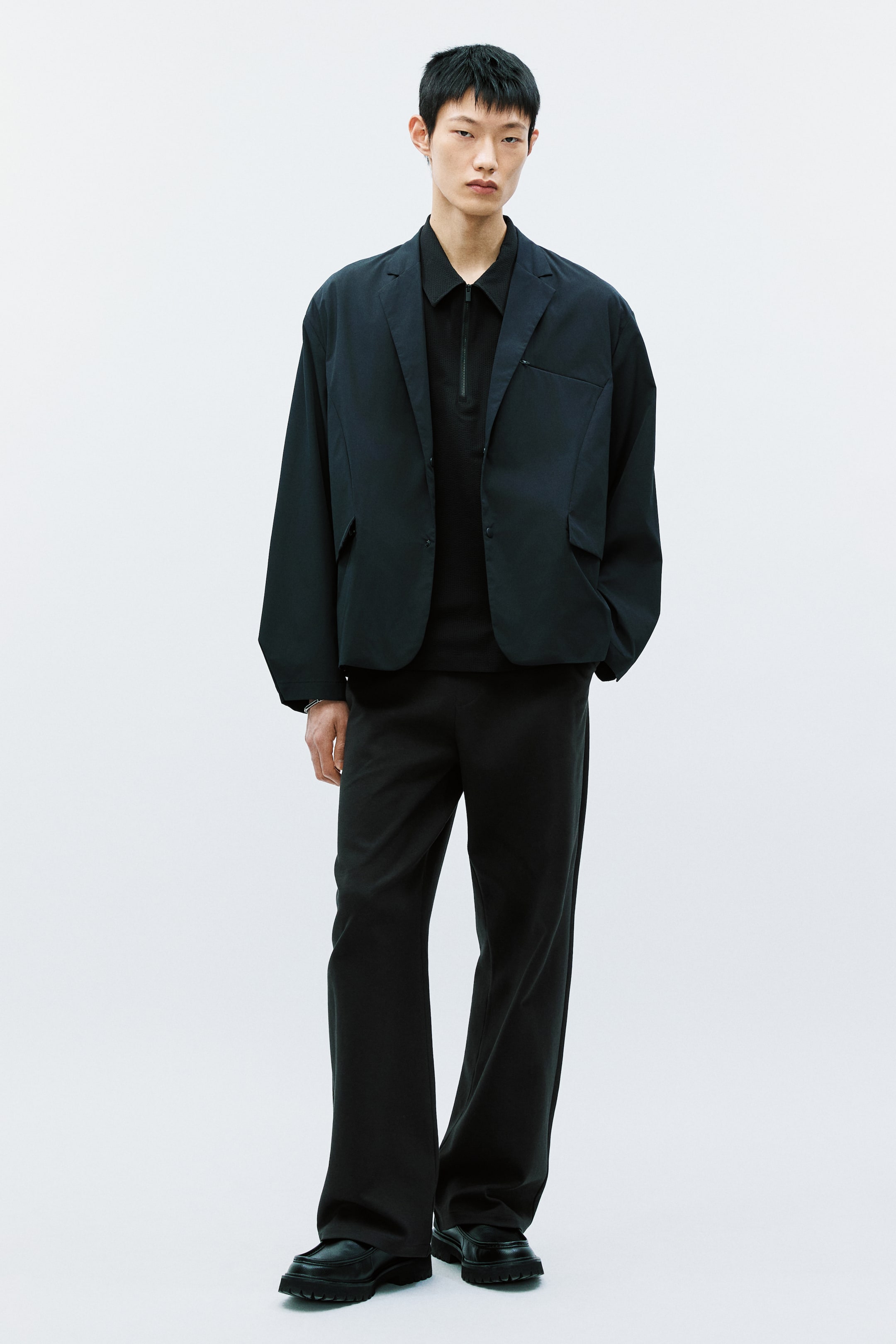 이미지 확대 보기: 어두운 색상의 싱글 브레스티드 블레이저는 오픈 스타일로 착용되었으며, 노치 라펠, 가슴 패치 포켓, 두 개의 하단 패치 포켓이 특징입니다. 이 블레이저는 반지퍼 클로저가 있는 리브드 다크 폴로 셔츠 위에 레이어드 되었으며, 어두운 와이드 레그 트라우저와 어두운 러그 솔 로퍼와 매치되었습니다.