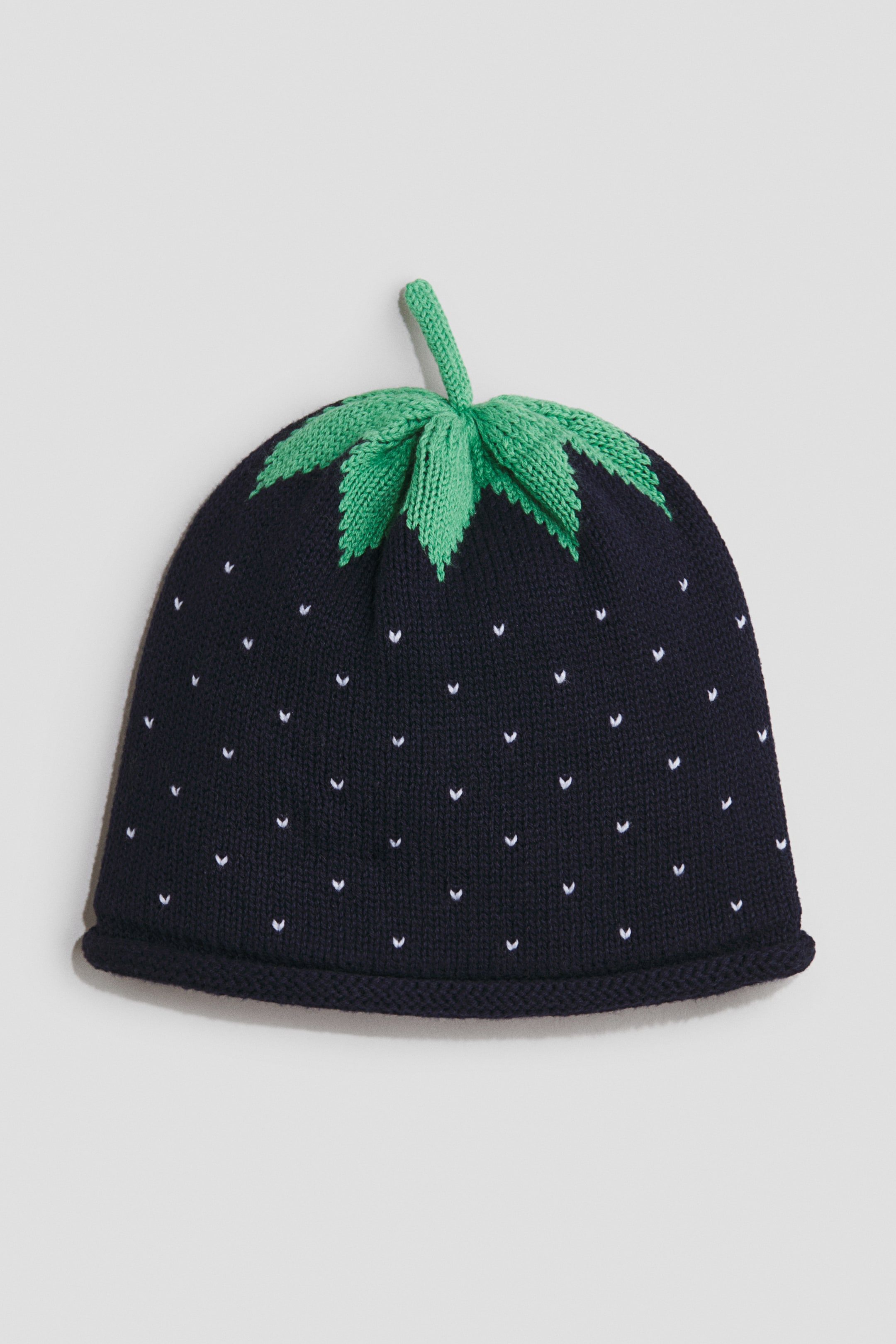 View larger image: A dark blue jacquard-knit cotton beanie is facing forward, showcasing small white V-shaped dots across its surface. A green knitted stem and leaves create a strawberry-like detail on its top, while a rolled-up edge completes the design at the bottom.
