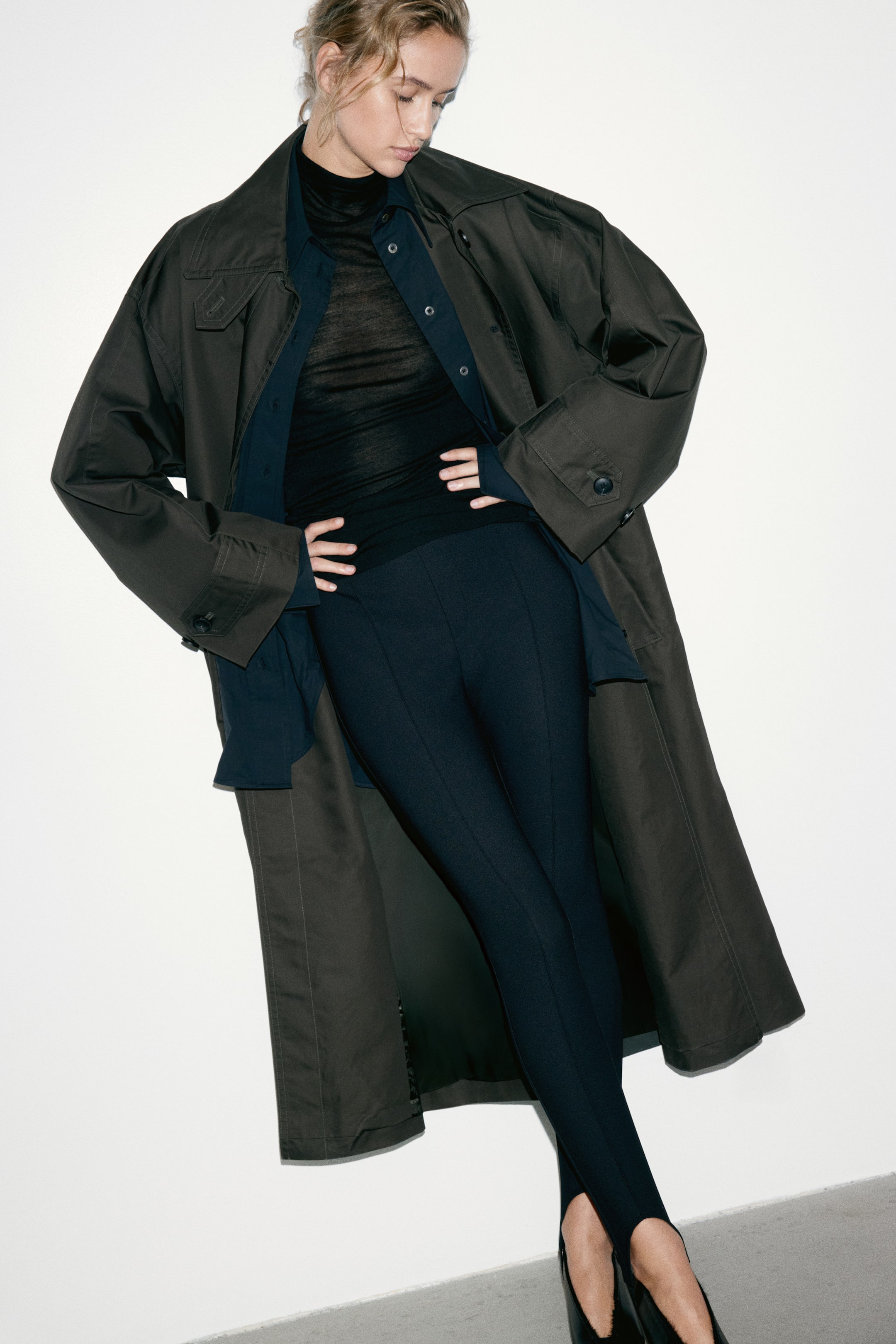 View larger image: The Crease Front Stirrup Leggings in dark navy are being worn by the model, featuring a sharp vertical seam down the centre of each leg and stirrup straps looped under the feet. The leggings are styled with a layered black high-neck top, a navy blue button-down shirt, an oversized olive trench coat, and black high-heeled shoes.