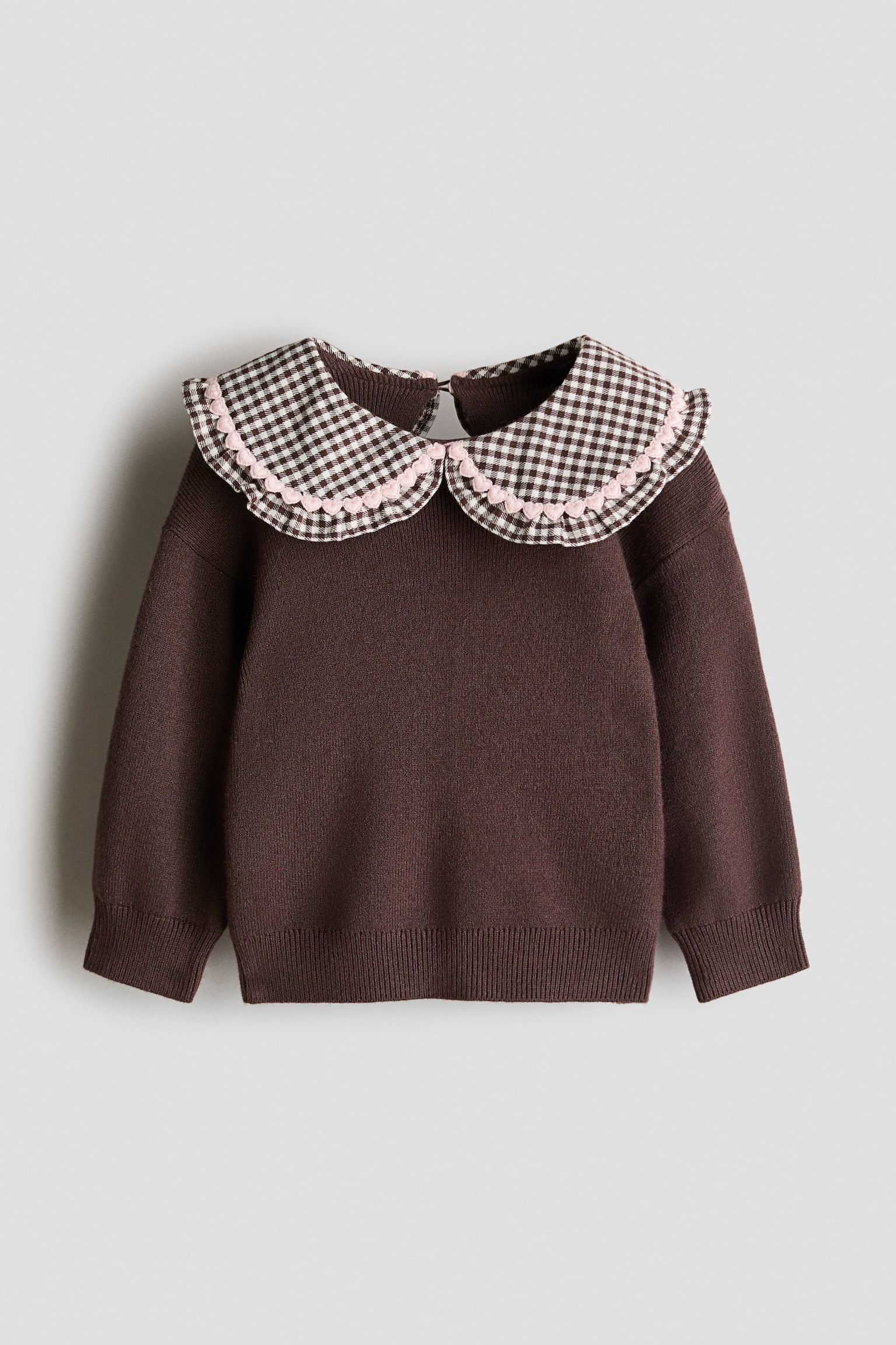 A brown knit jumper is shown from the front, featuring a large Peter Pan collar in a brown and white gingham pattern with a pink scalloped trim. The jumper has long sleeves with ribbed cuffs and a matching ribbed hem.