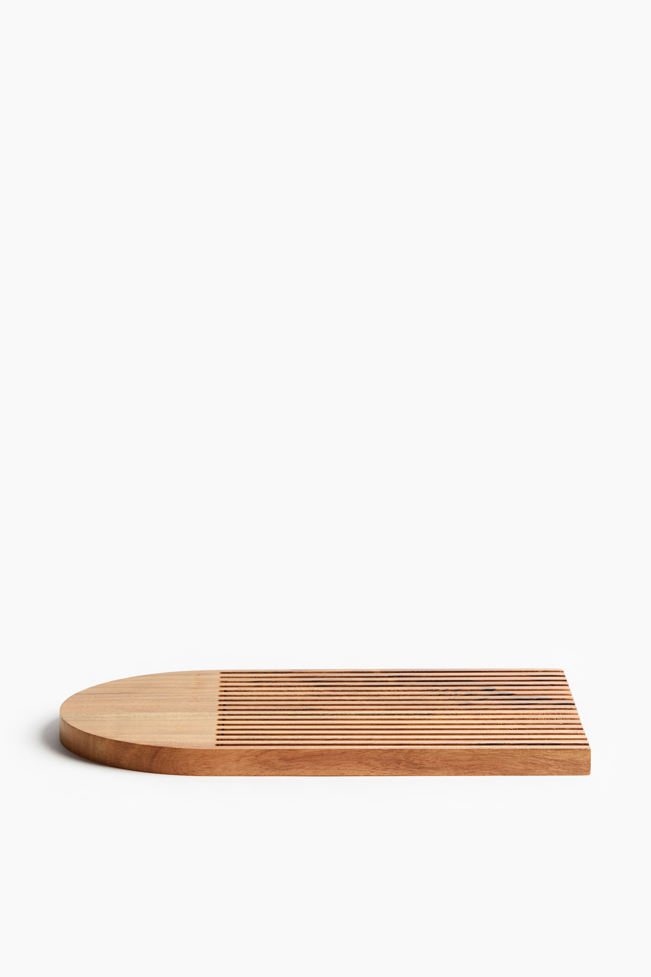 View larger image: An acacia wood chopping board is exhibiting a rectangular shape with one rounded end. The board showcases parallel grooves covering approximately two-thirds of its length from the straight end, with the rounded section remaining smooth.