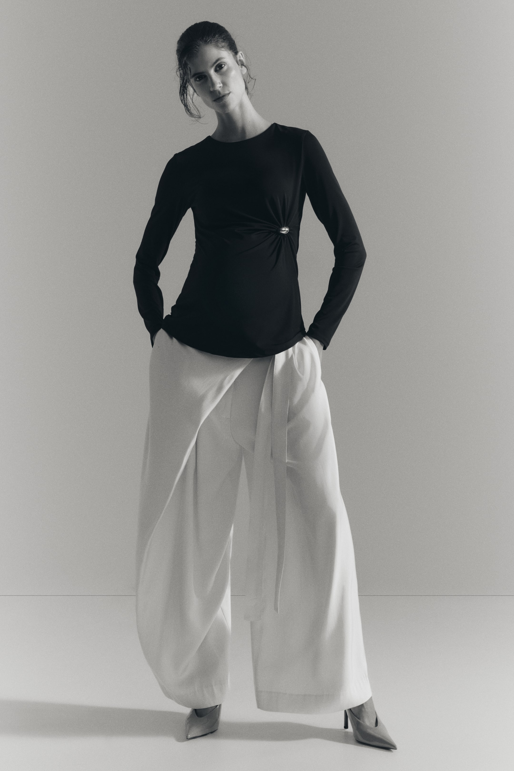 View larger image: The model is wearing a fitted black jersey top with a round neckline and long sleeves, featuring a circular metal detail on one side that gathers the fabric to create a draped effect. The top is styled with light-coloured, wide-leg trousers.