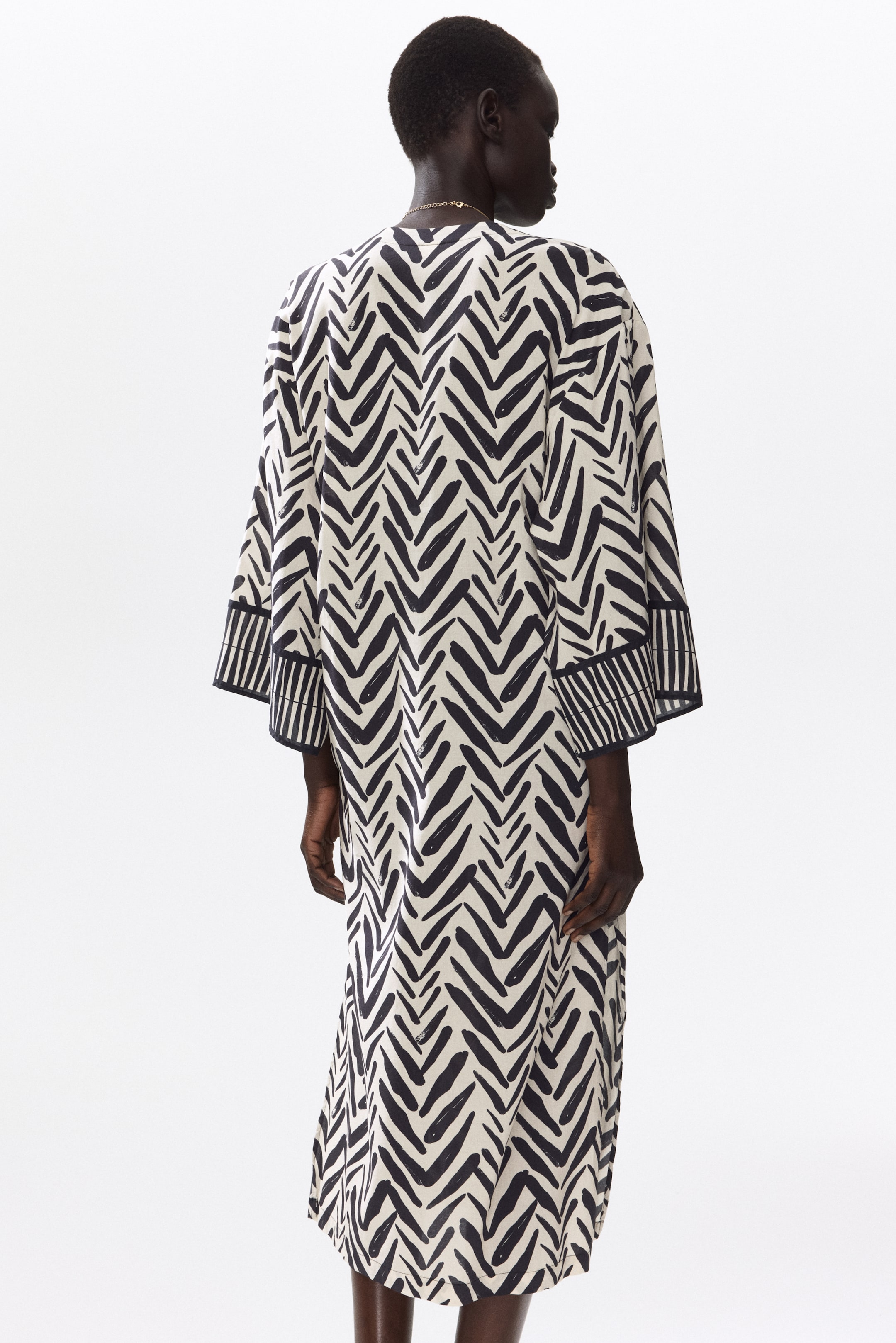 View larger image: The model is wearing a calf-length tunic dress featuring a black and white chevron pattern. It showcases dropped shoulders and 3/4-length sleeves accented with striped cuffs. Around the model's neck is a delicate gold necklace.