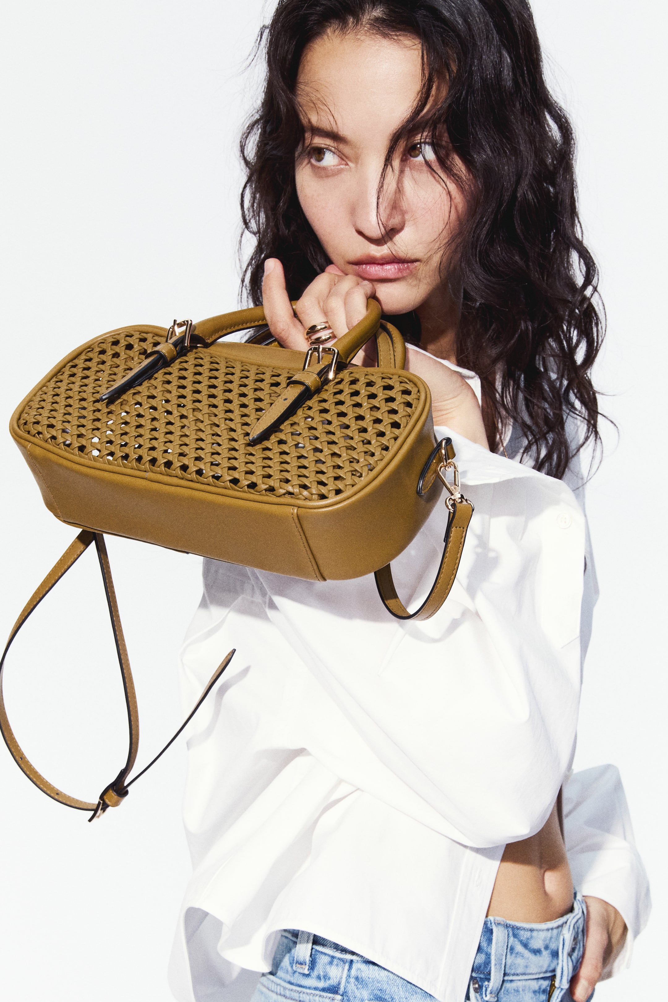 查看大圖: 模特手持一款卡其色斜挎包，採用塗層面料製成。包包正面設計有開放式格紋編織，頂部手柄可調節並配有金屬扣，還附有一條可拆卸、可調節的肩帶垂掛。