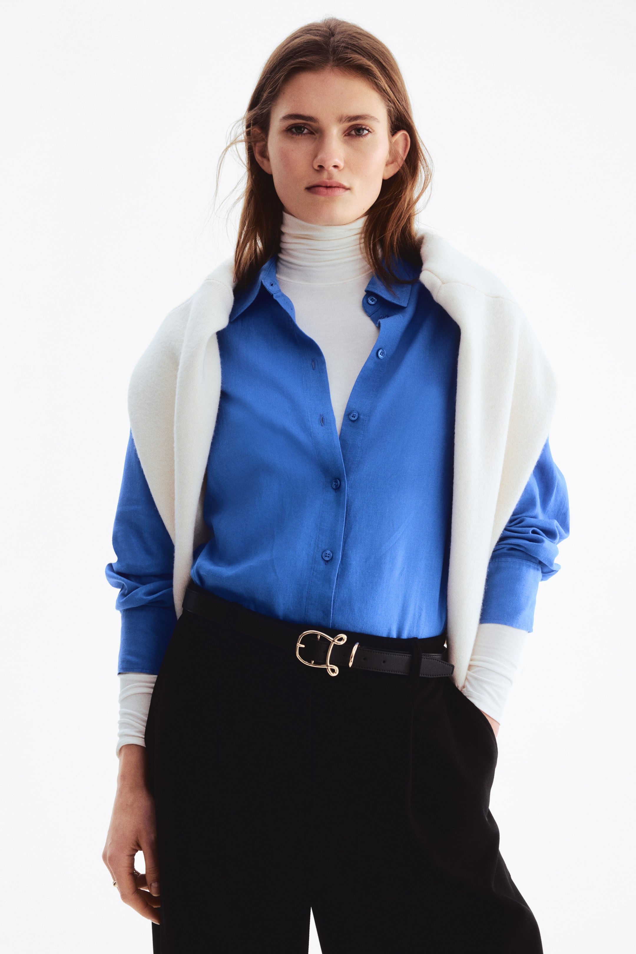 View larger image: A blue button-front shirt is styled open over a white turtleneck, complemented by a white sweater elegantly draped across the shoulders. It showcases a pointed collar and long sleeves, casually pushed up at the wrists.