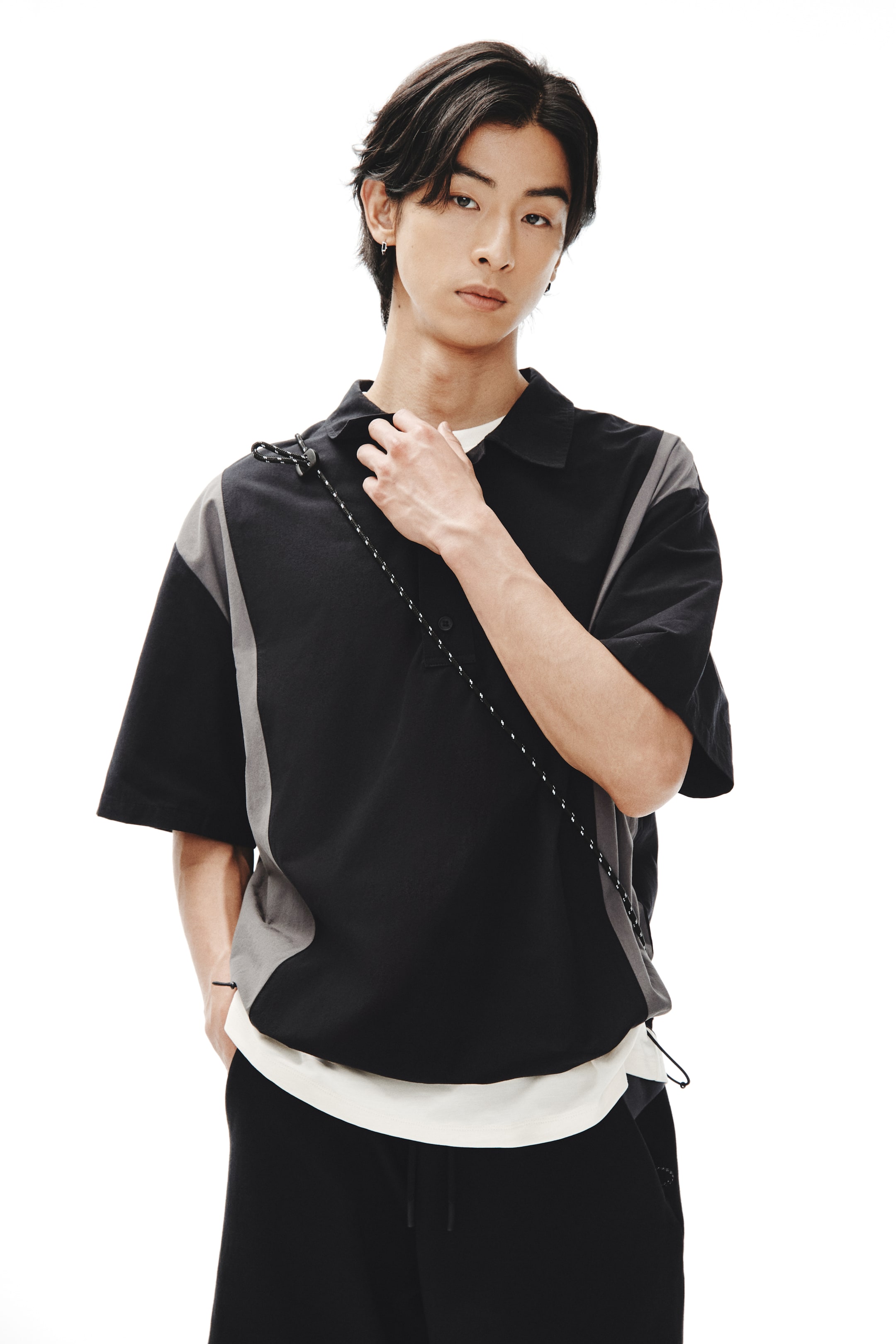 Xem ảnh lớn hơn: Người mẫu đang mặc một chiếc áo sơ mi popover bằng cotton màu đen và xám với cổ áo, hàng cúc, tay ngắn và các tấm bên màu xám. Chiếc áo có dây rút đàn hồi với chốt dây ở gấu, tạo nên một dáng vẻ rộng rãi, và người mẫu đang điều chỉnh cổ áo bằng một tay, để lộ ra một chiếc áo lót màu sáng.