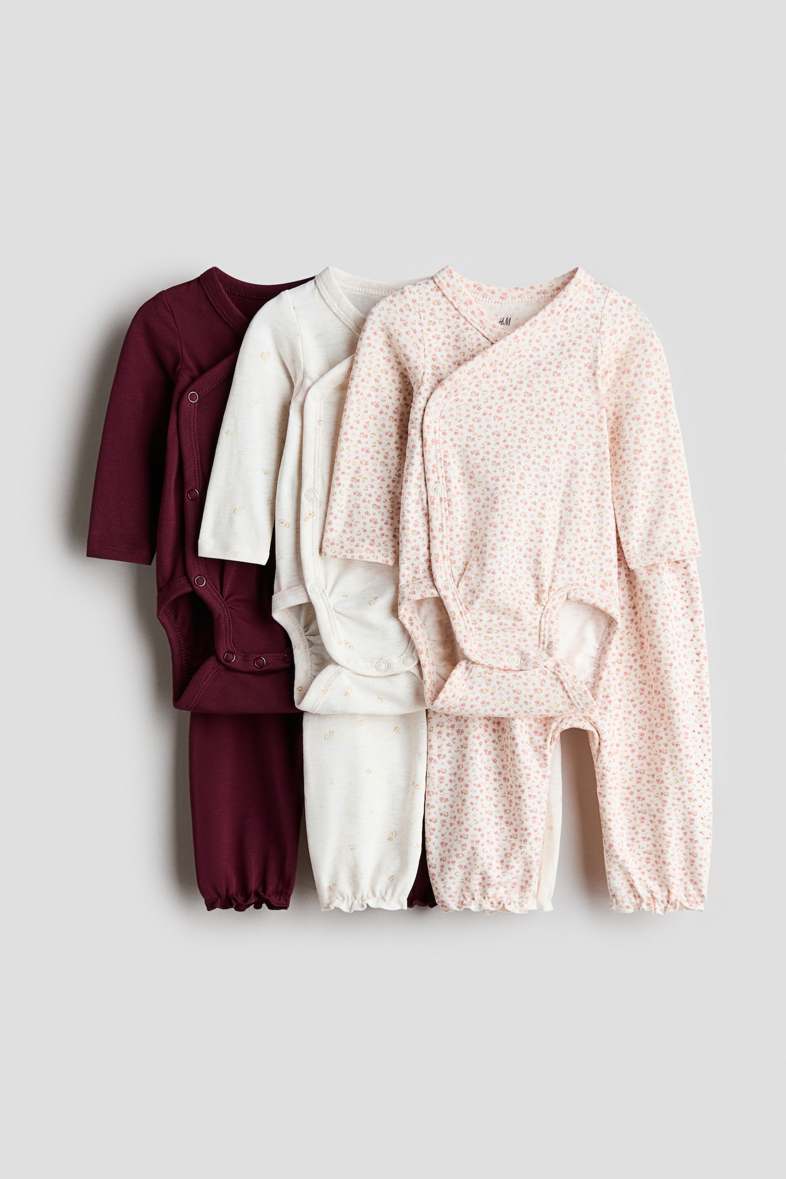 Three baby outfits are hanging facing front, with each set displaying a long-sleeve, wrap-front bodysuit and matching trousers. The sets are shown in solid burgundy, patterned cream, and pink floral print, with the bodysuits featuring side popper fastenings and the trousers showing gathered cuffs.